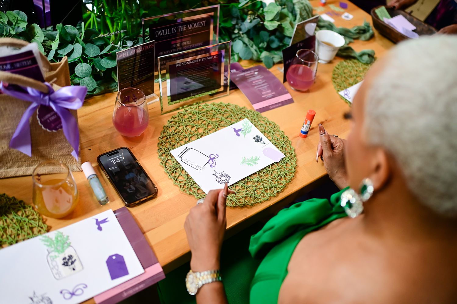 Woman in green dress writing on paper at decorated event table with drinks and floral arrangements.