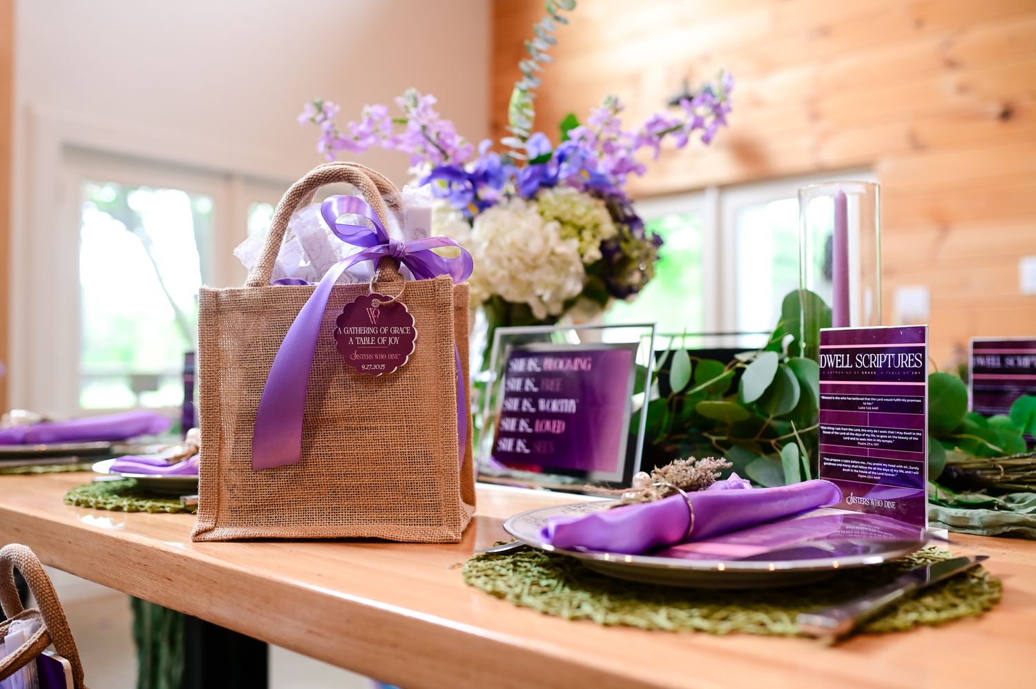 Elegant purple wedding table setting with burlap gift bag, floral centerpiece, and purple napkins on moss placemats.