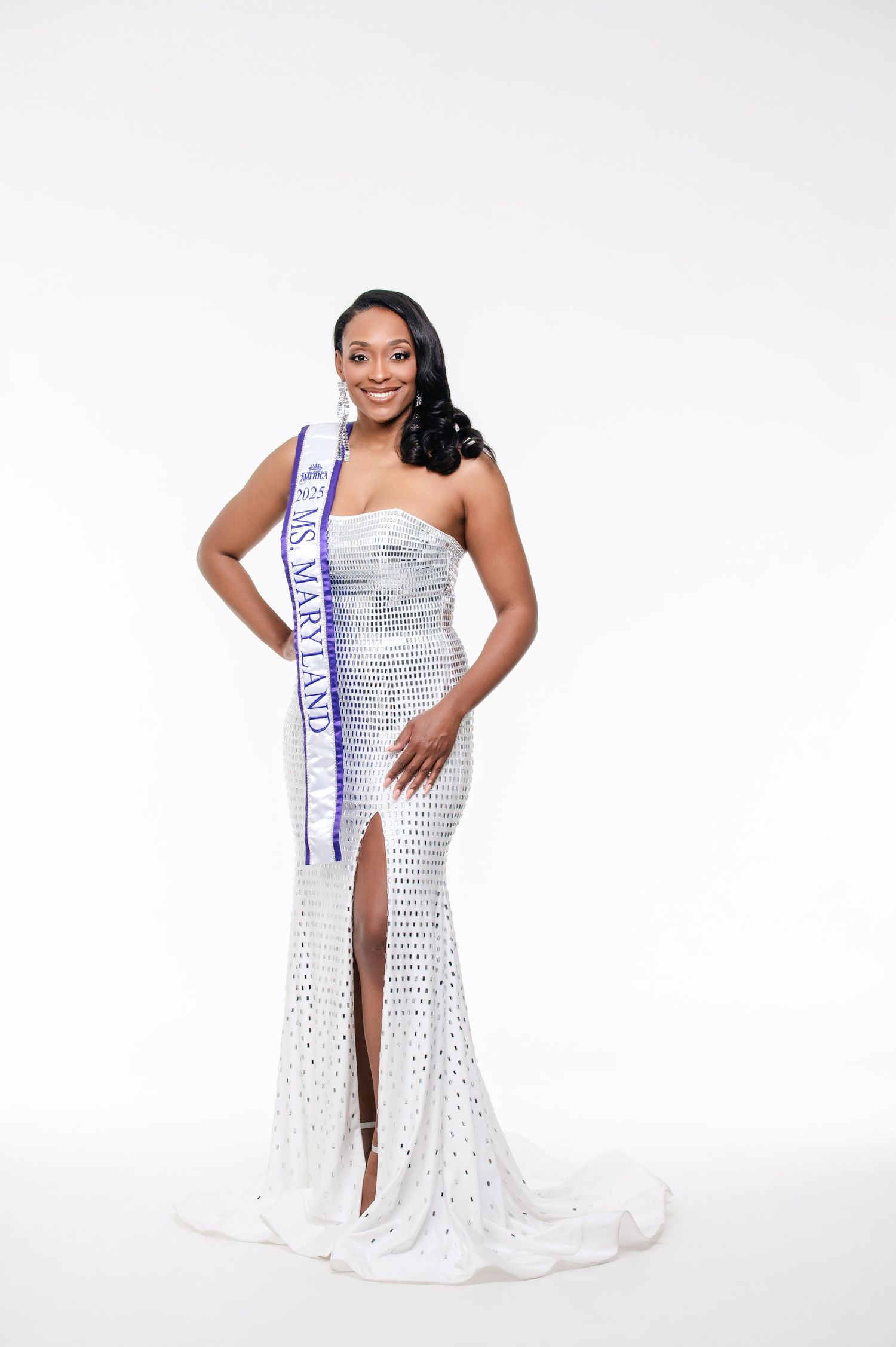 A pageant contestant poses in a sparkling white evening gown with a high slit against a white backdrop.