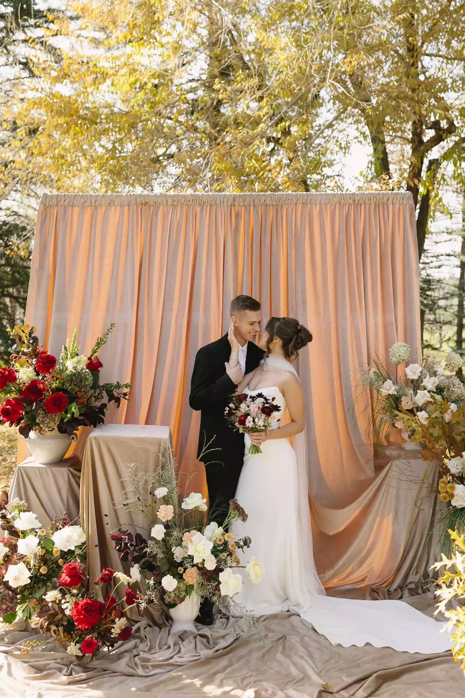 Wedding couple poses against a blush pink backdrop with red and white floral arrangements under autumn trees.