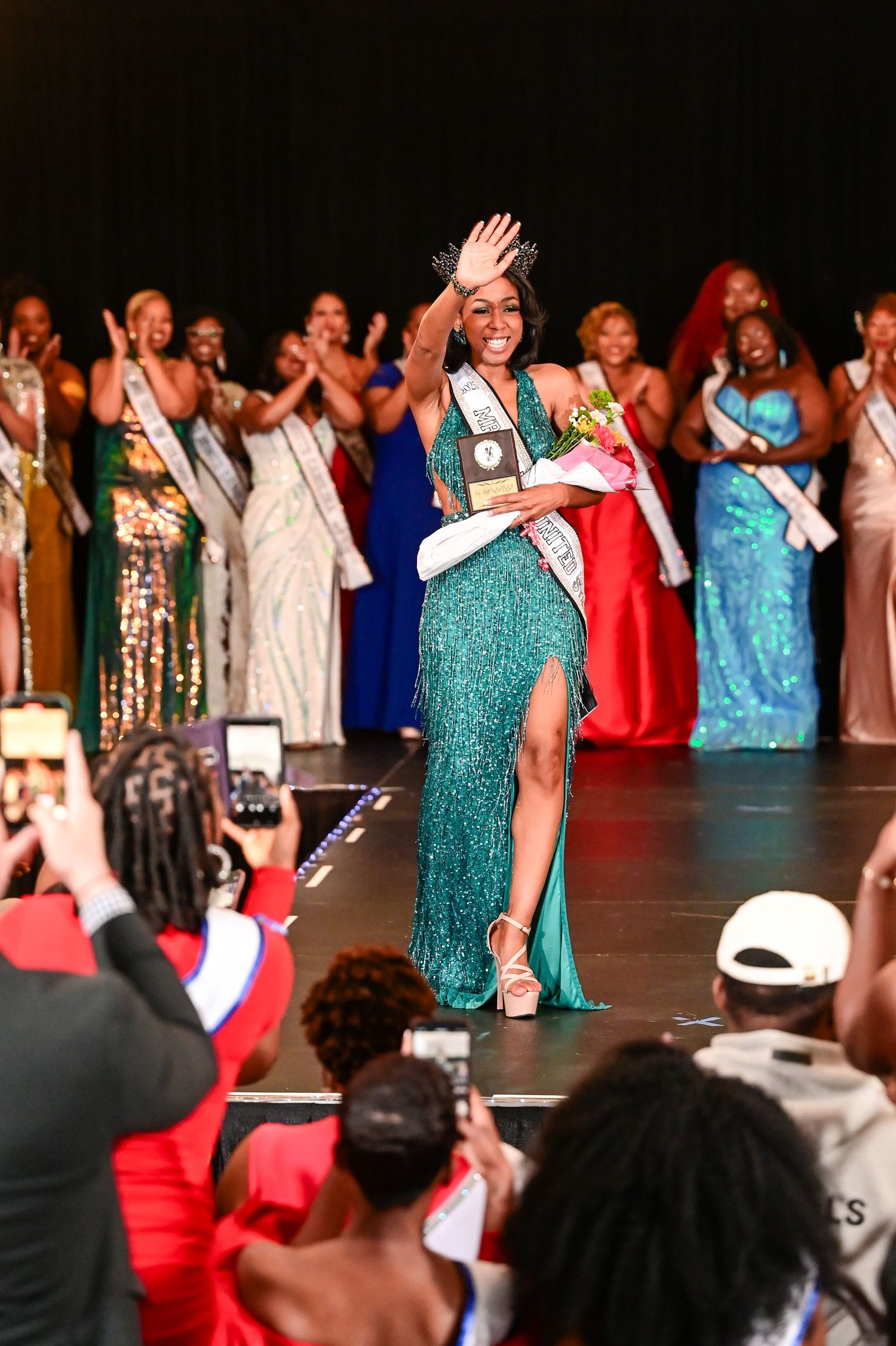 A group of performers in colorful dresses and outfits stand on stage with their hands raised during a show.
