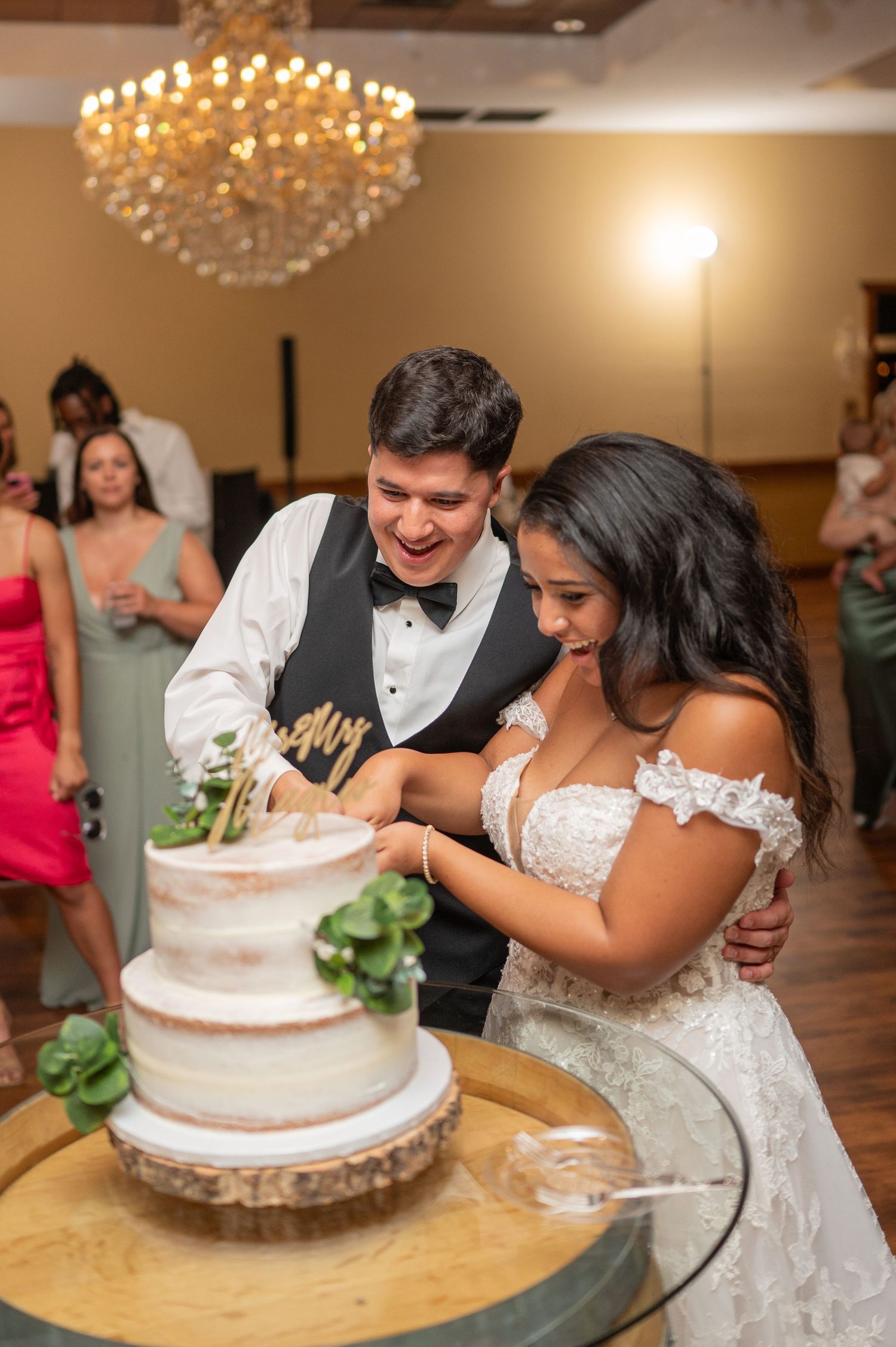 A couple shares a joyful moment cutting their semi-naked wedding cake under a crystal chandelier at their reception.