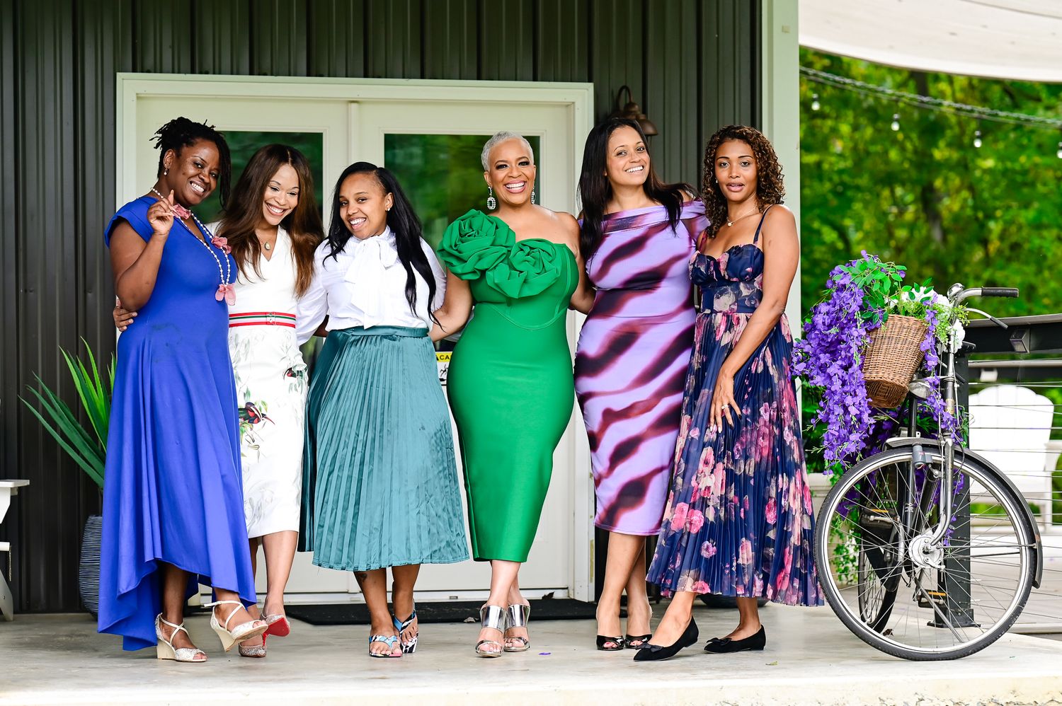 Six stylish women posing together outdoors in colorful dresses, smiling in front of a building with a bicycle nearby.