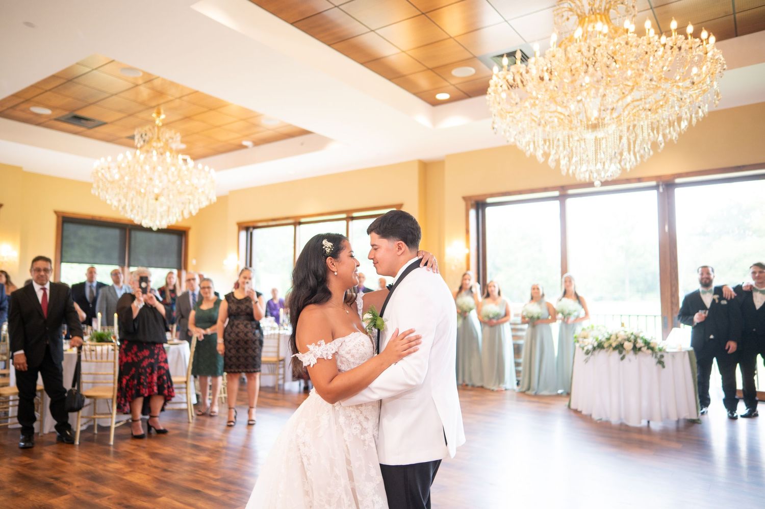 A couple shares their first dance in an elegant ballroom with crystal chandeliers and large windows.
