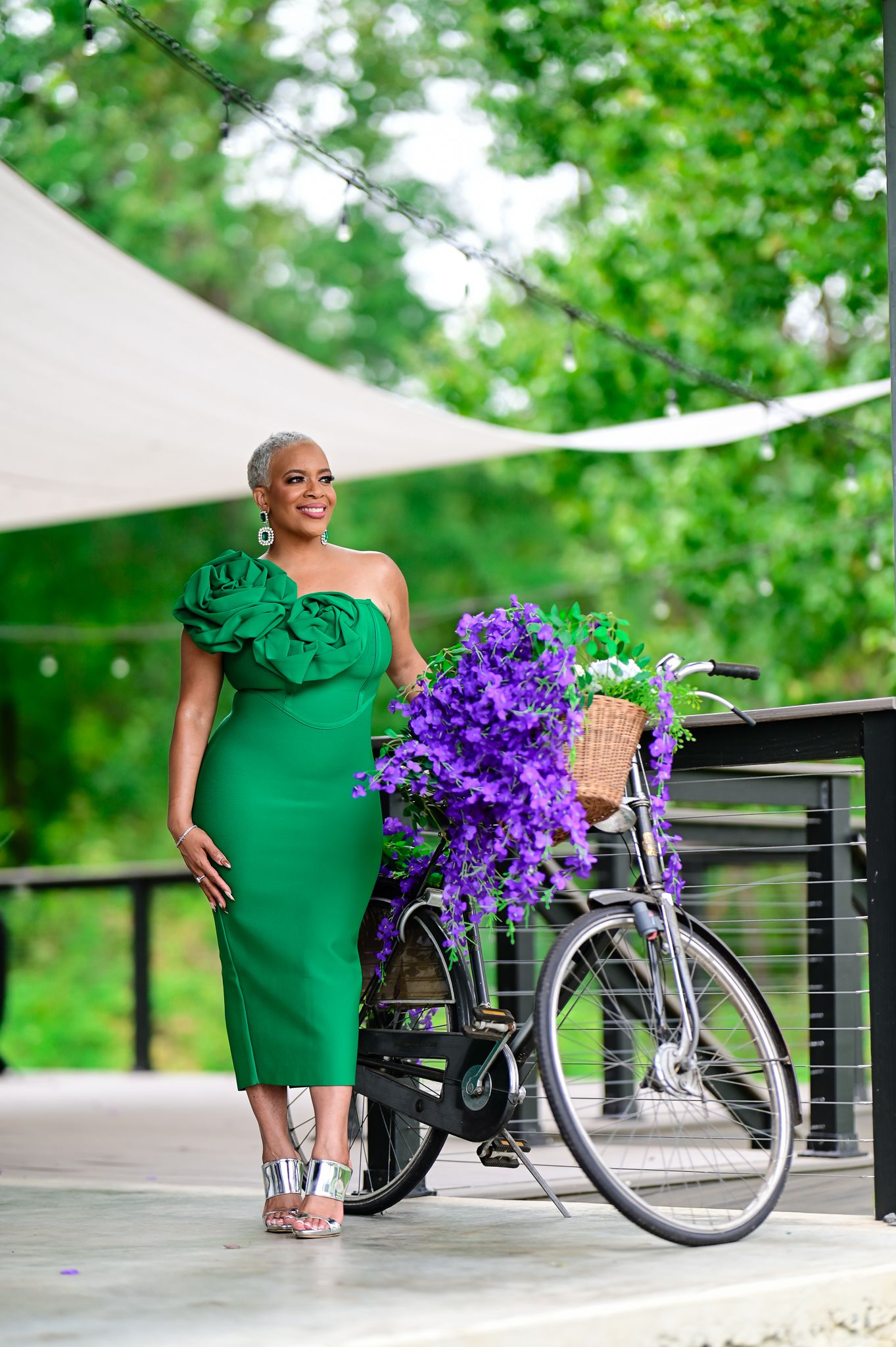 Elegant woman wearing a green ruffle dress standing next to a flower-decorated bicycle on a scenic outdoor deck.