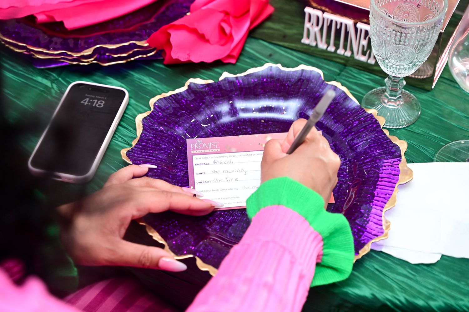 Someone wearing pink and green writes on paper with a purple decorative plate and smartphone on a green tablecloth.