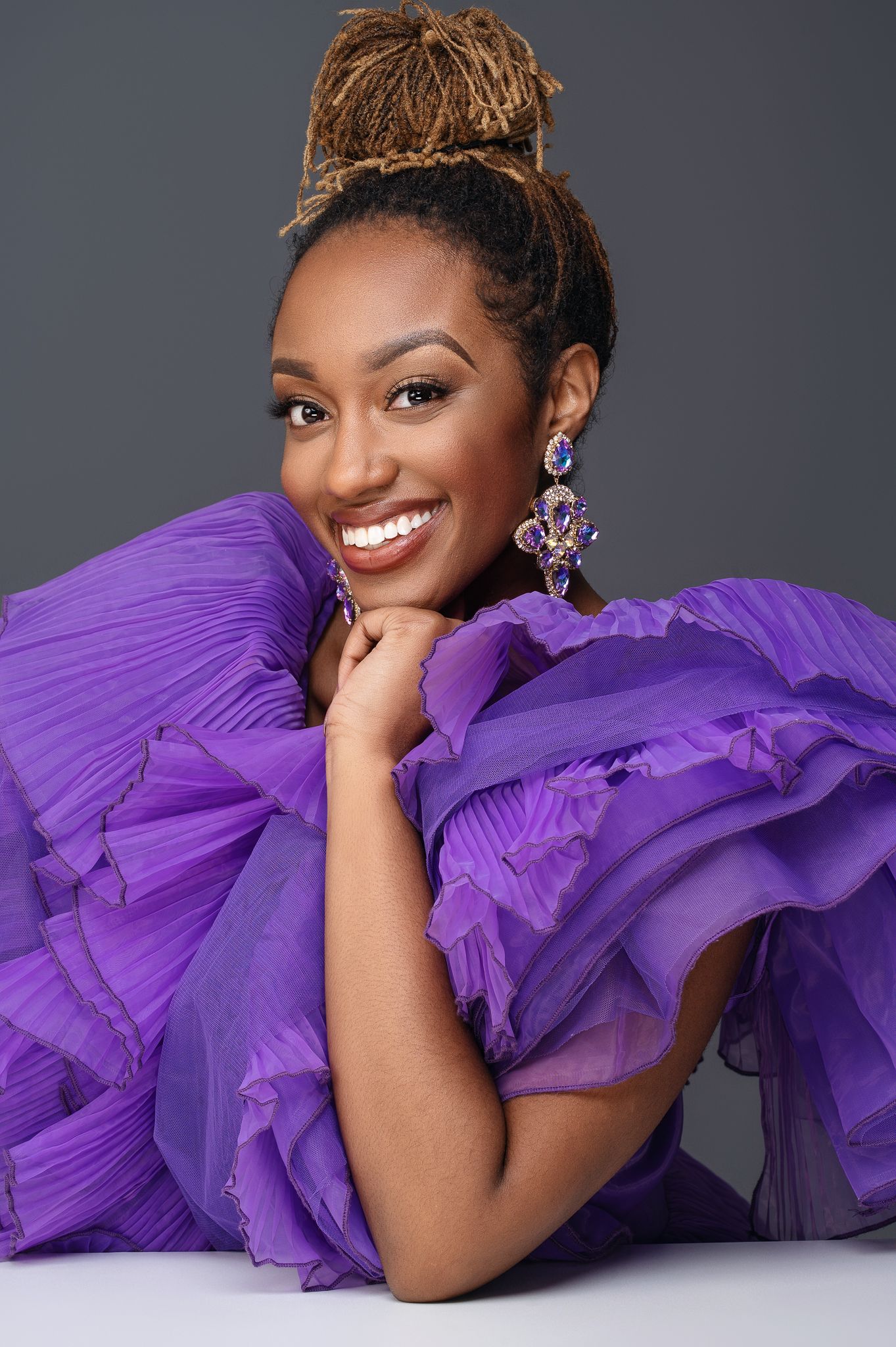 Elegant portrait in violet ruffled gown with statement earrings against gray background.