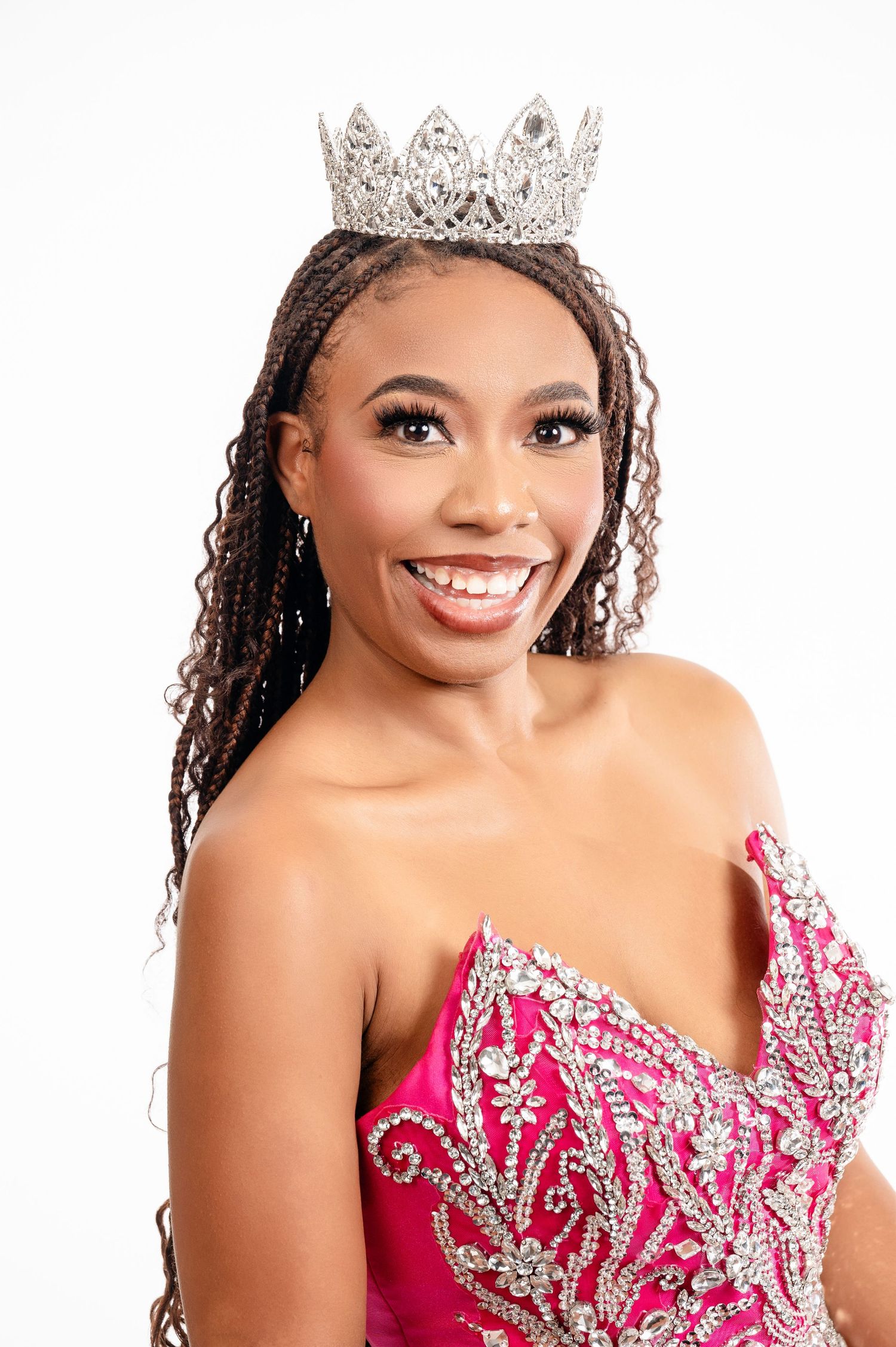 A pageant contestant wearing a silver crown and sparkling pink gown with beaded embellishments smiles at the camera.