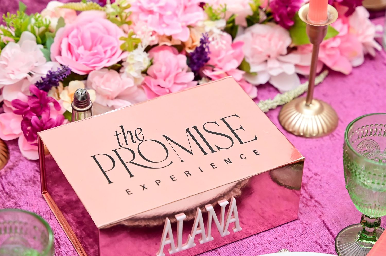 Pink floral arrangement surrounds an elegant rose gold box labeled 'The Promise Experience' on a magenta tablecloth.
