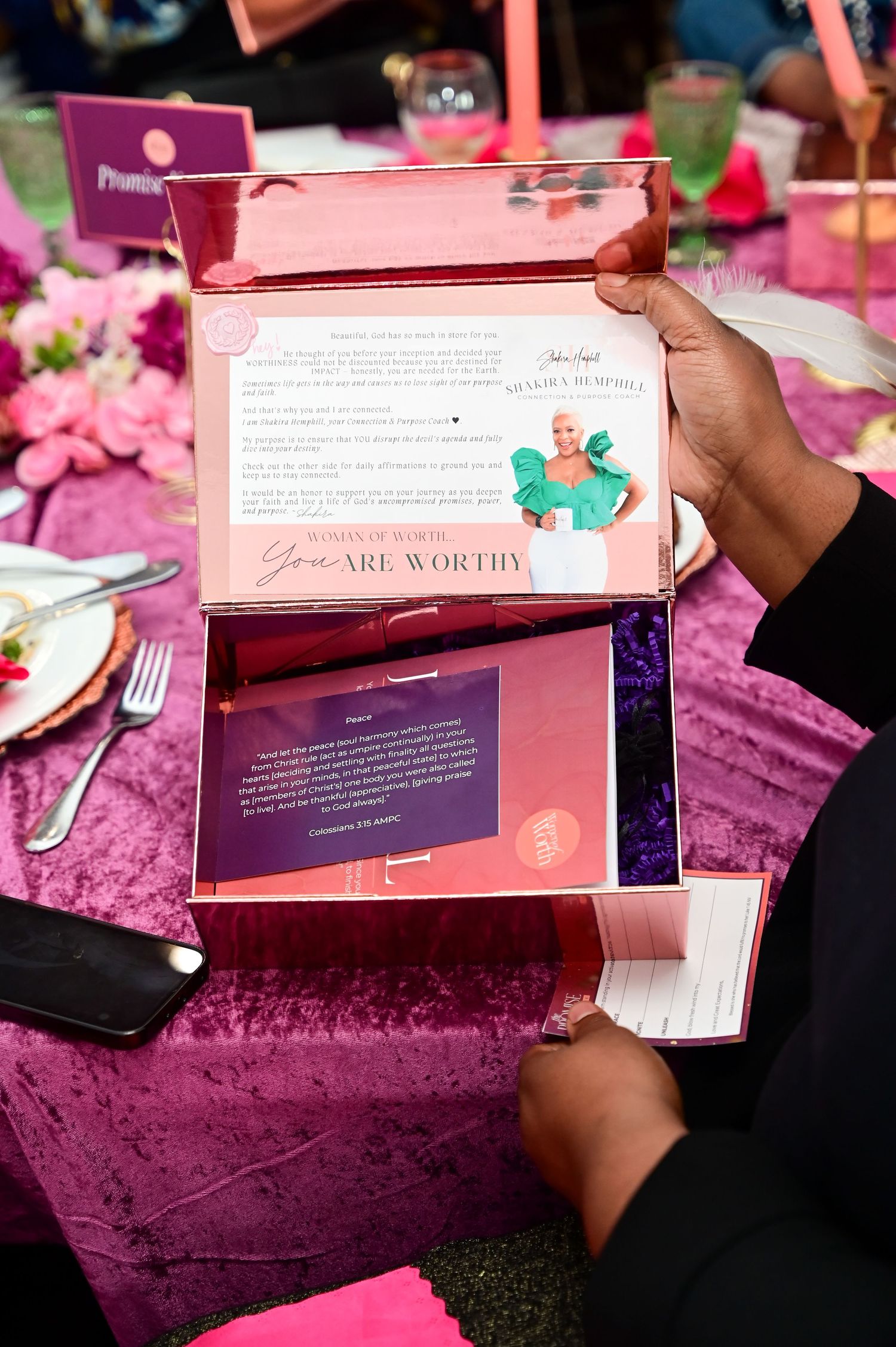 Hands opening a pink gift box with inspirational message cards on a purple velvet tablecloth at an elegant event setting.