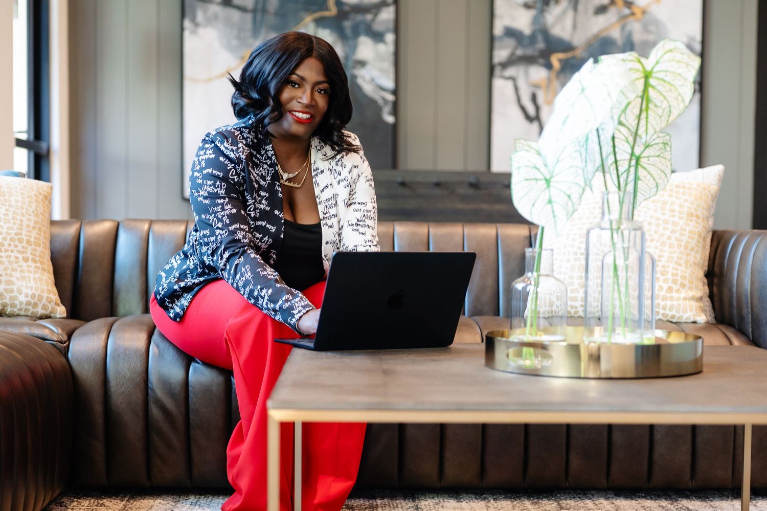 Professional woman in red pants and blazer smiling while working on laptop in modern lounge setting.