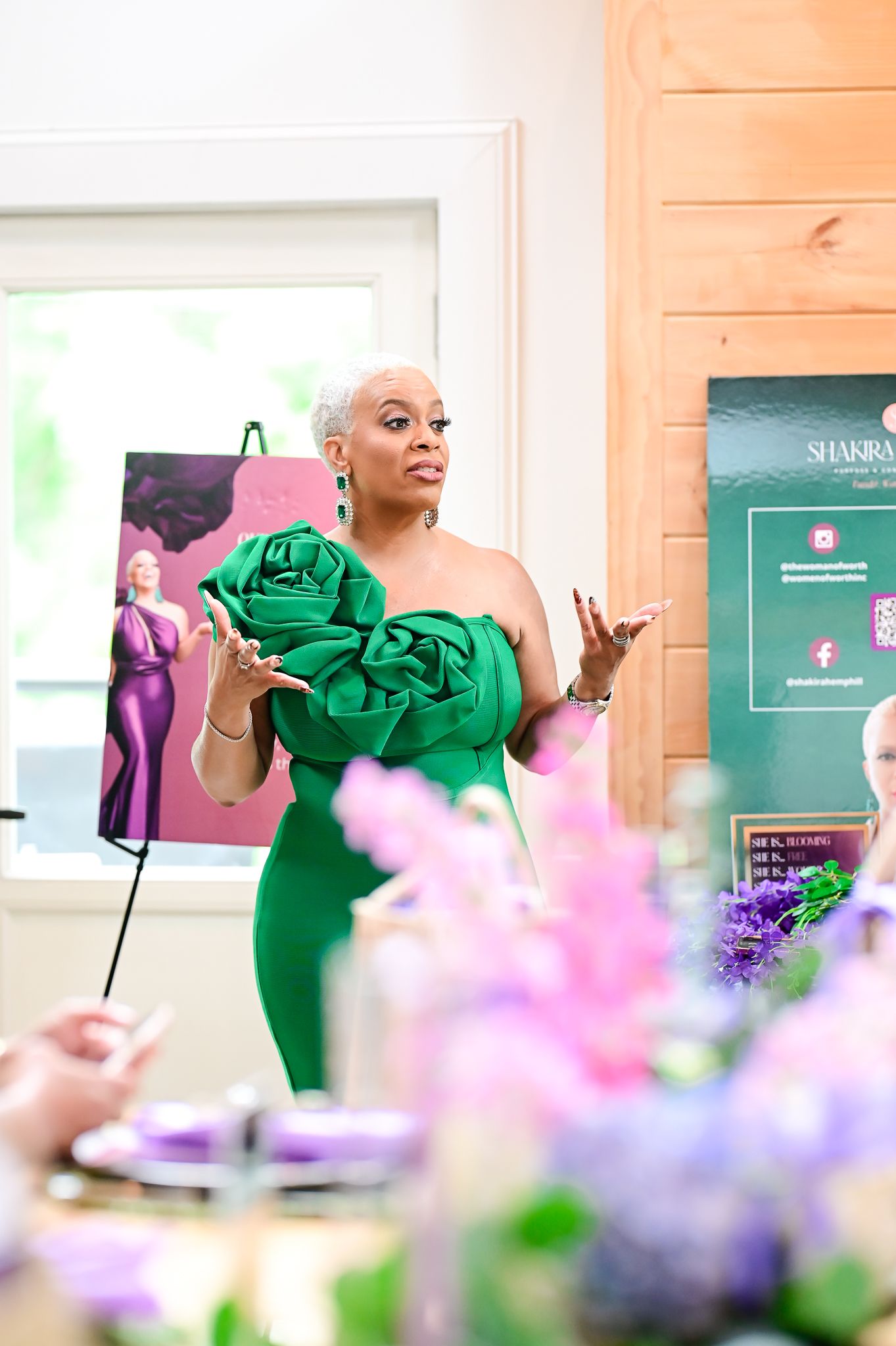 Woman in elegant green off-shoulder gown with rose detail speaks at an event with floral decorations.