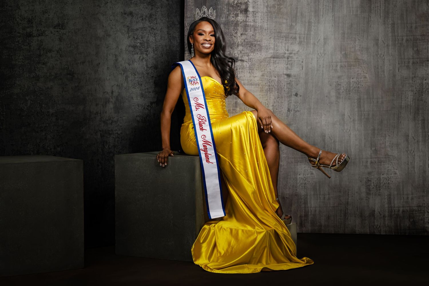 Woman in yellow gown wearing pageant sash and crown poses elegantly in professional portrait photo.