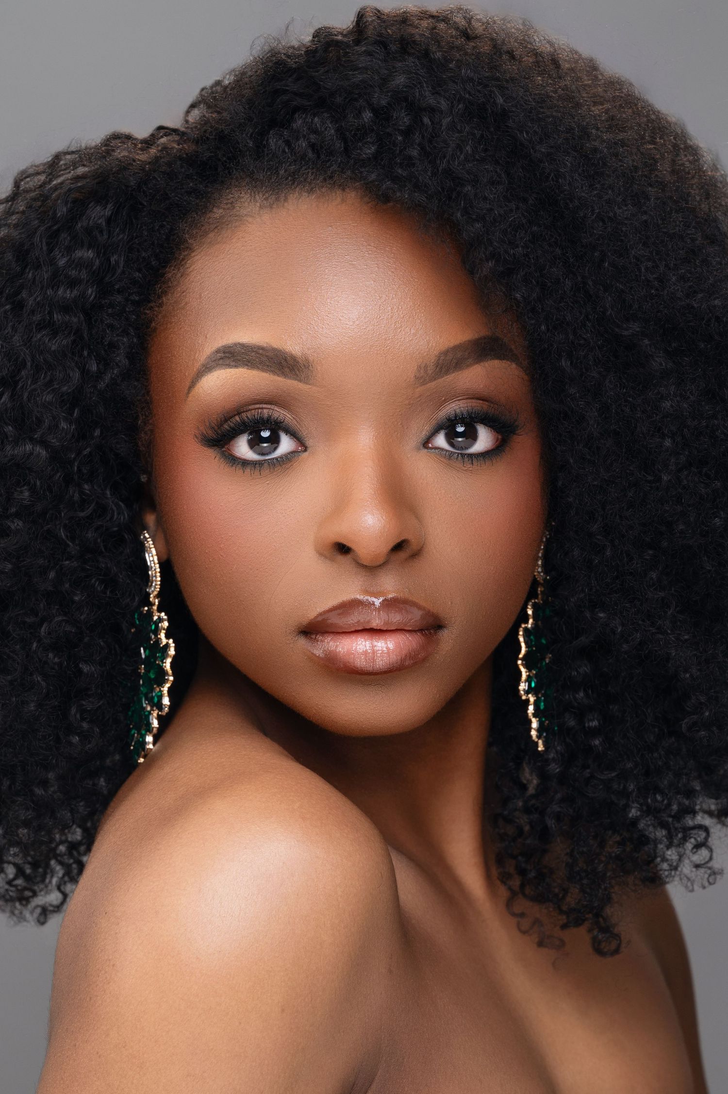 Beauty portrait of a woman with natural curly afro hair and elegant makeup wearing dangling earrings against gray backdrop.