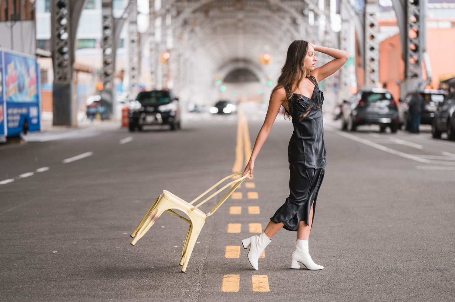 A woman in a black dress and white boots dances in a city tunnel with a golden chair on a city street.