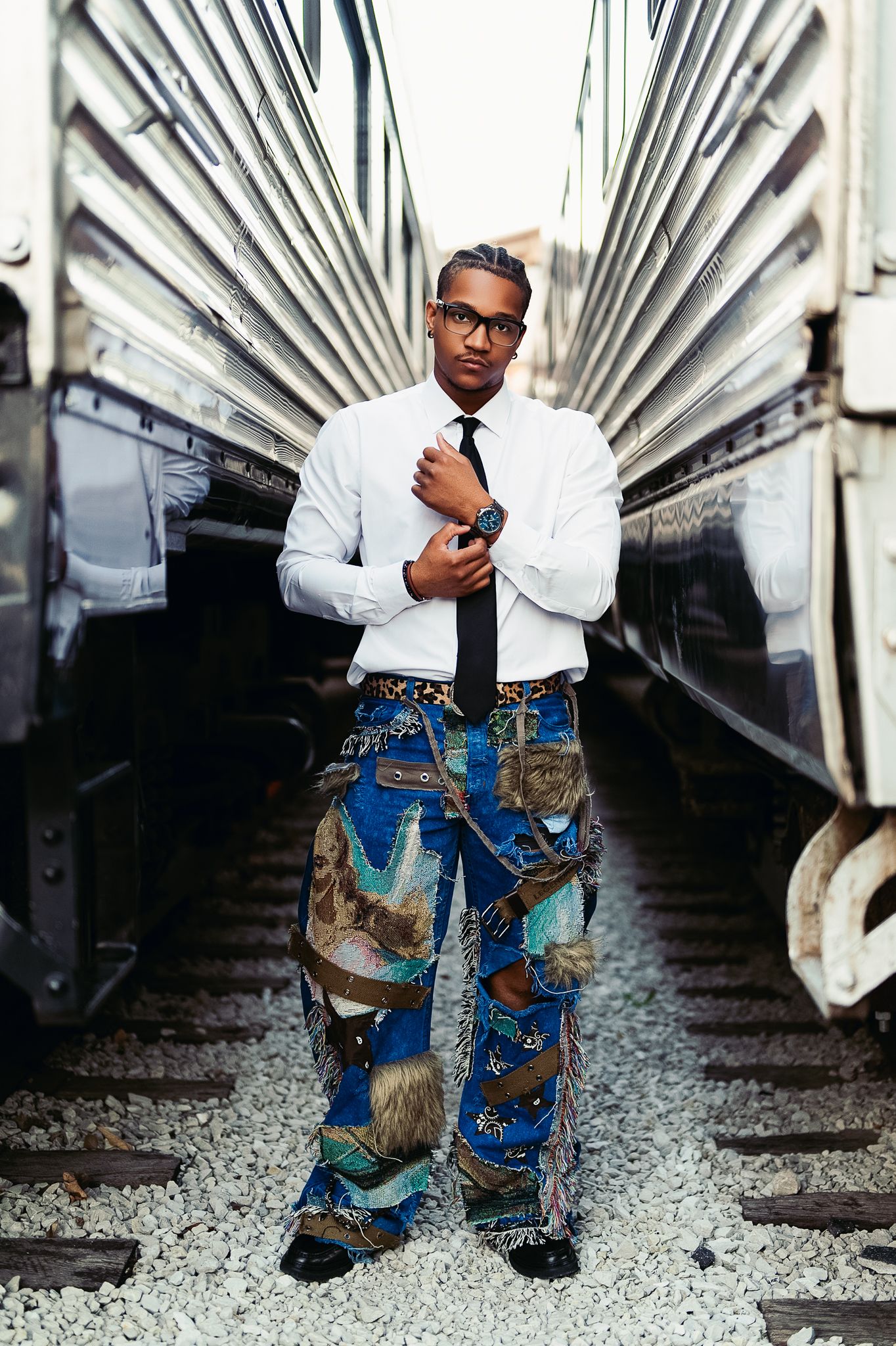 Stylish man in white shirt, black tie, and colorful patterned wide-leg jeans standing between train cars.