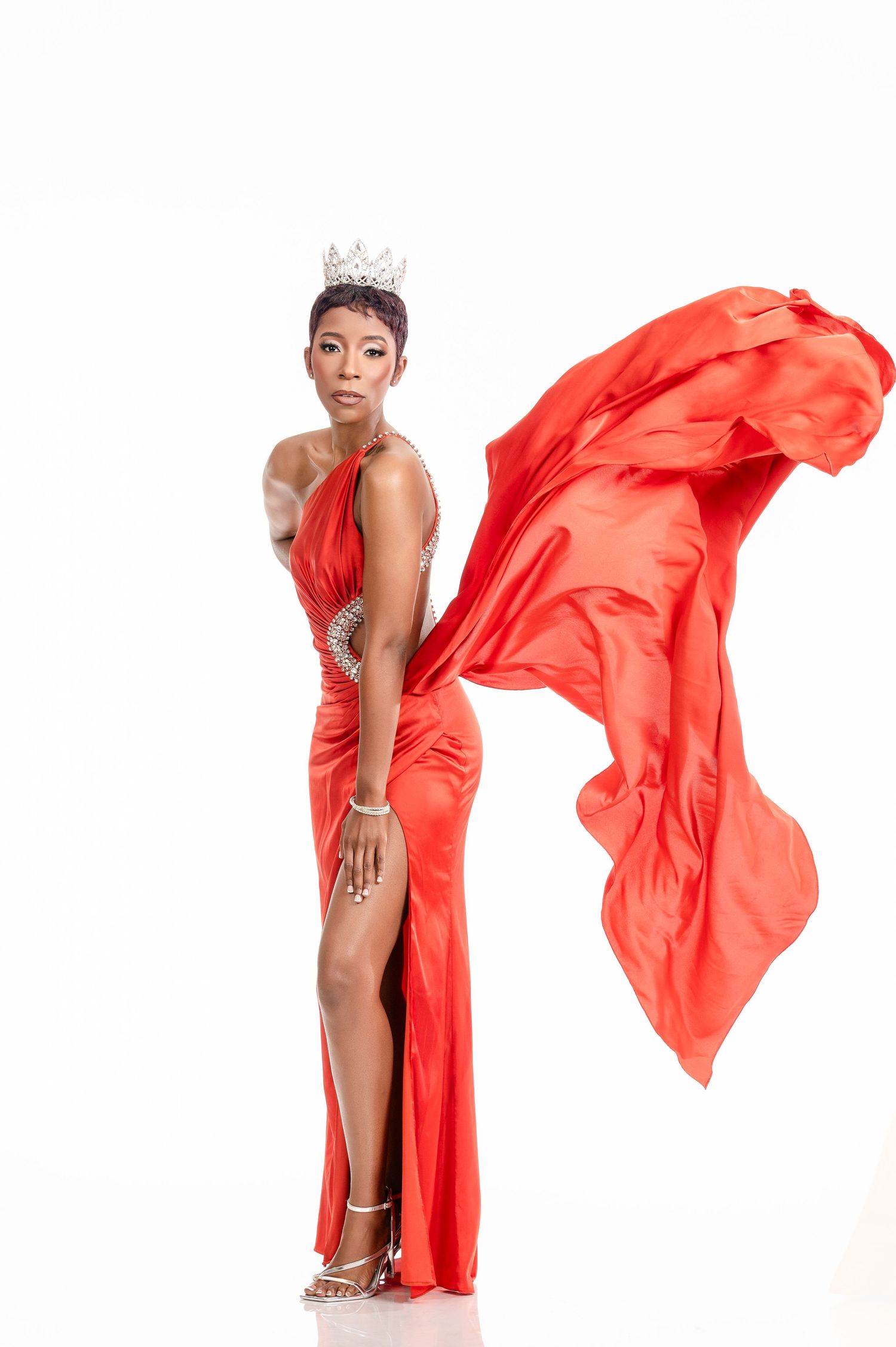Elegant model in flowing red gown with high slit and silver crown poses gracefully against white background.