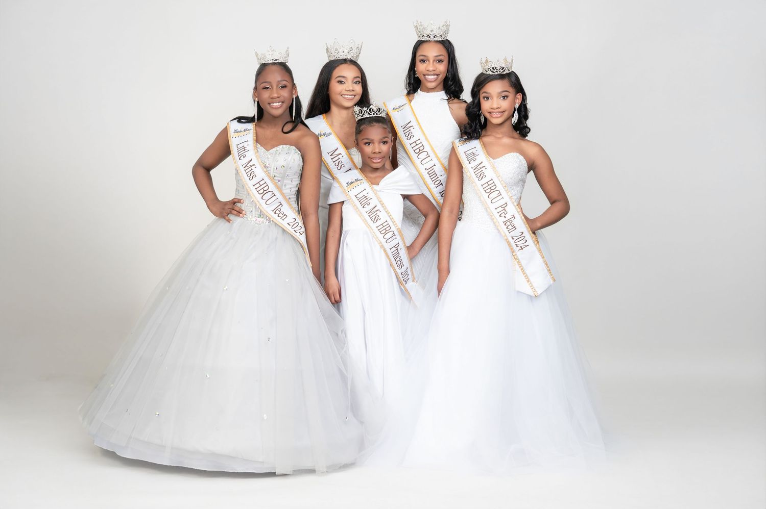 Five pageant contestants wearing white gowns and sashes pose together for a formal portrait against a light backdrop.