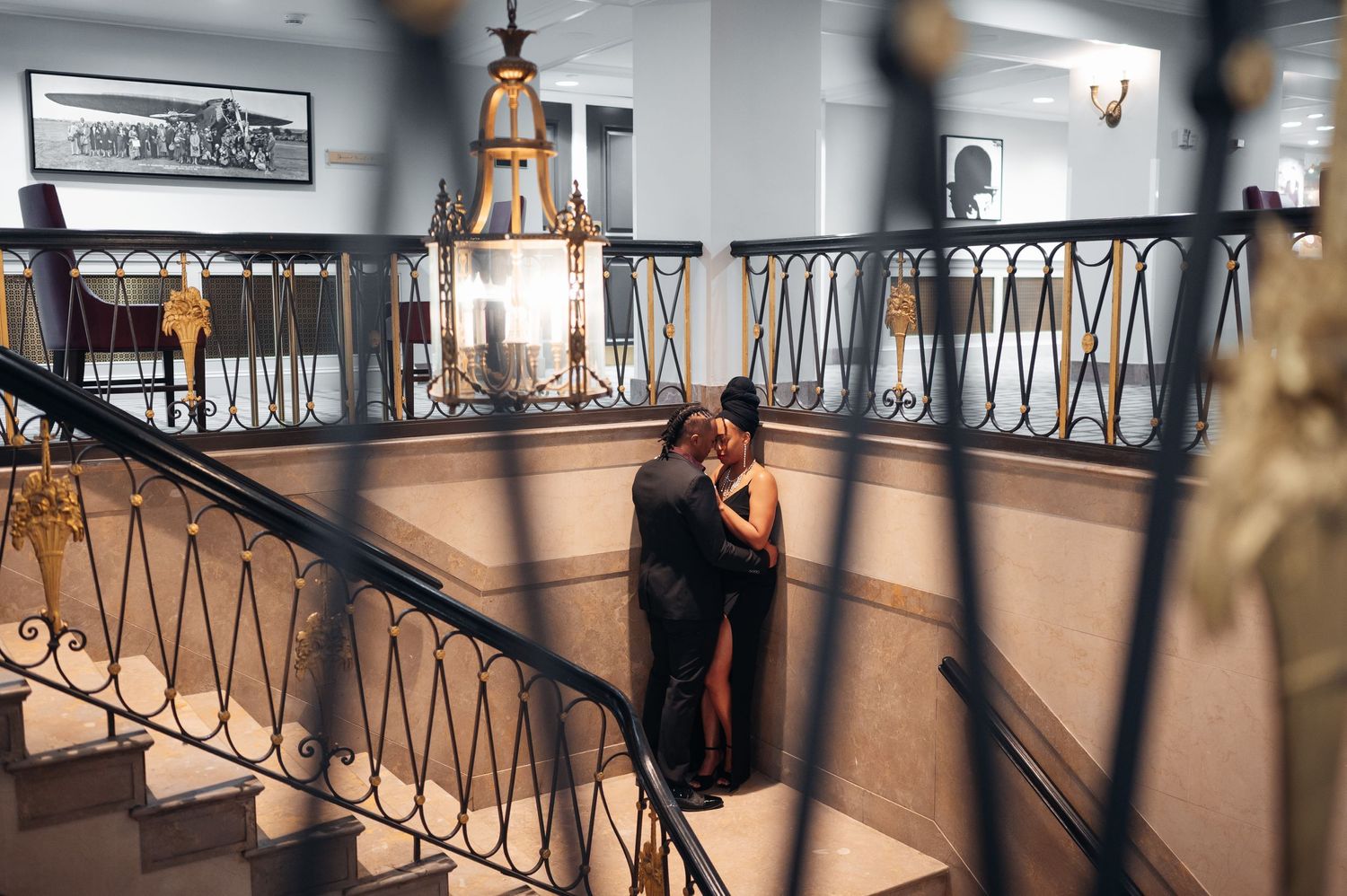 A couple embraces on an elegant interior balcony with wrought iron railings and chandelier lighting casting warm shadows.