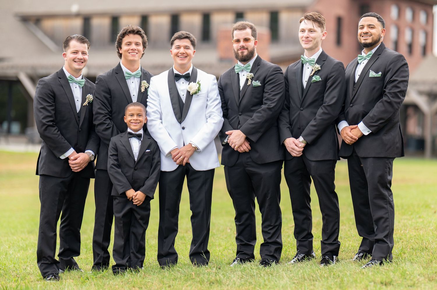 Wedding party in white and black formal attire posing together on a grassy lawn outside a brick building.