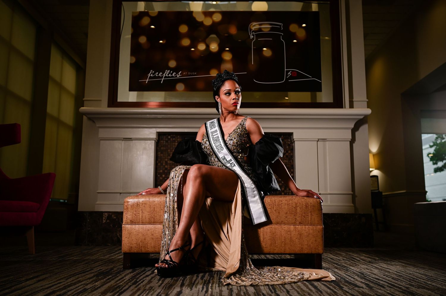 Beauty pageant contestant wearing a sash sits elegantly on brown leather furniture in an upscale room setting.