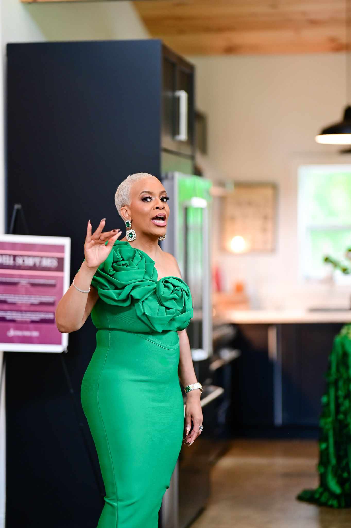 Woman in elegant green off-shoulder ruffle dress with matching earrings gesturing while speaking indoors.