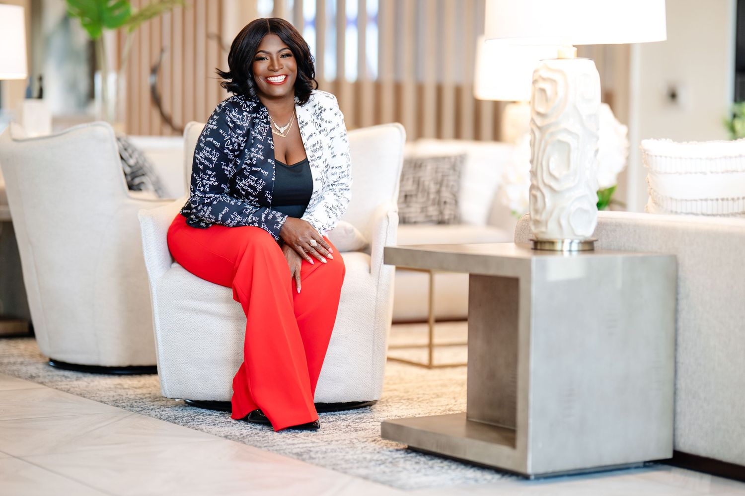 Confident Black woman in red pants and blazer sitting in modern white living room furniture.