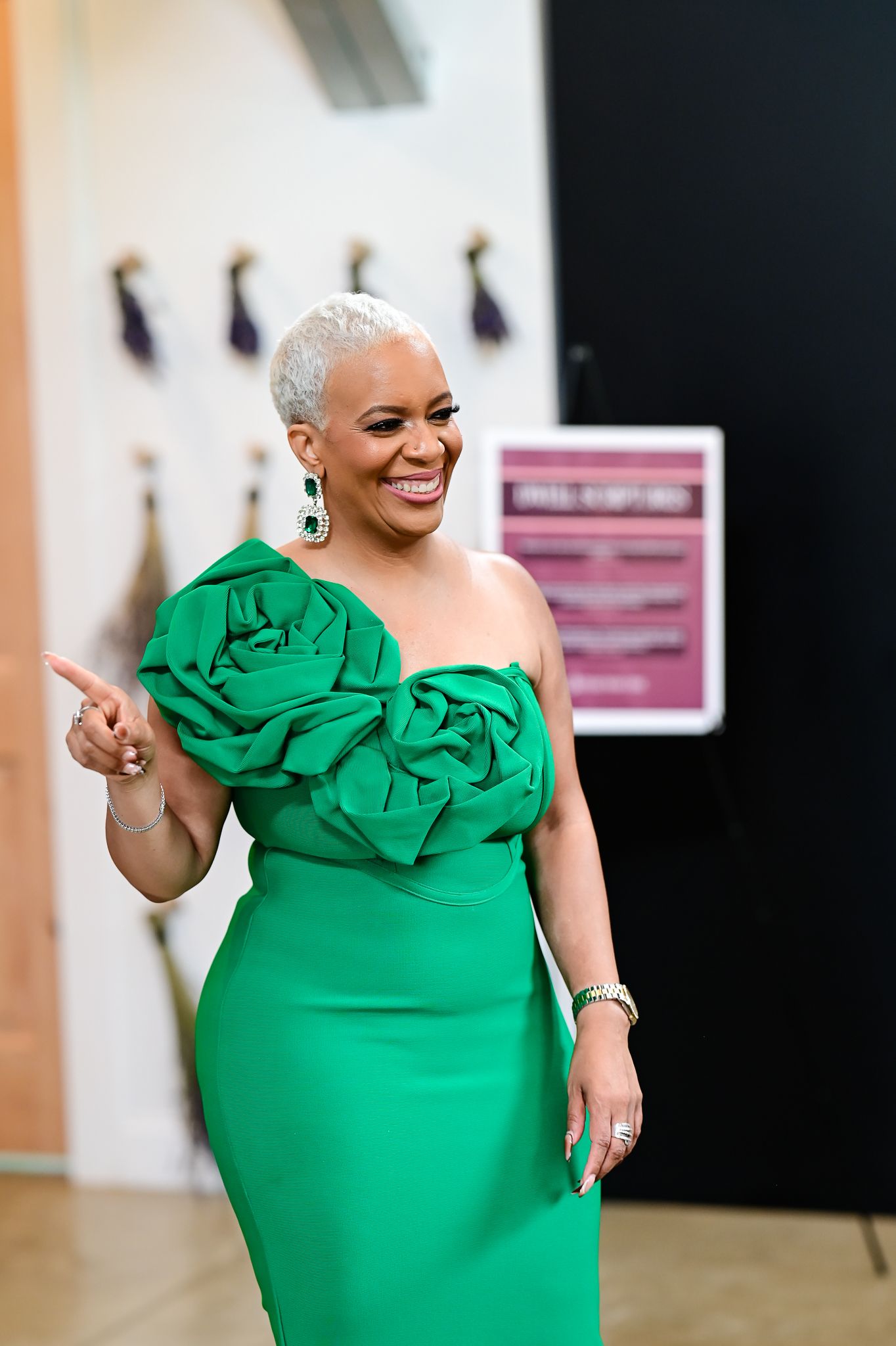 Woman in elegant green strapless rose applique gown with matching earrings, smiling and pointing at an event.