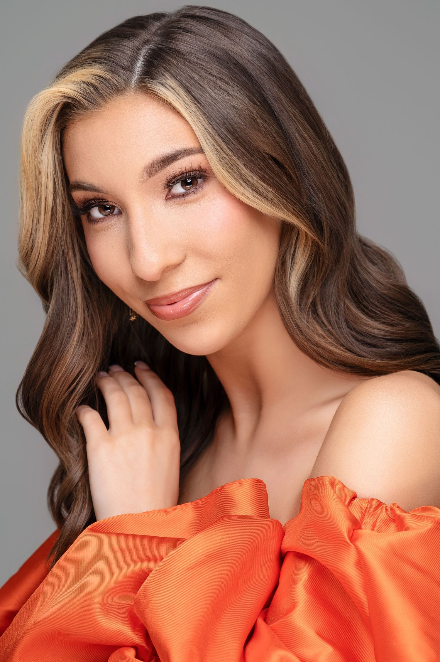 Studio portrait with warm lighting featuring a vibrant orange off-shoulder top and long wavy brown hair with highlights.