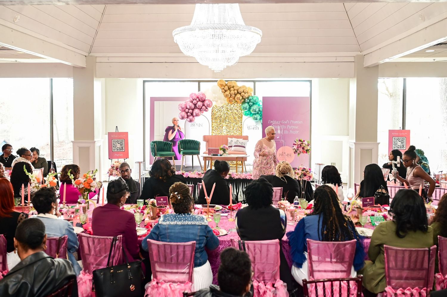 A decorated event space with pink chairs and colorful balloons features attendees gathered for a celebration.