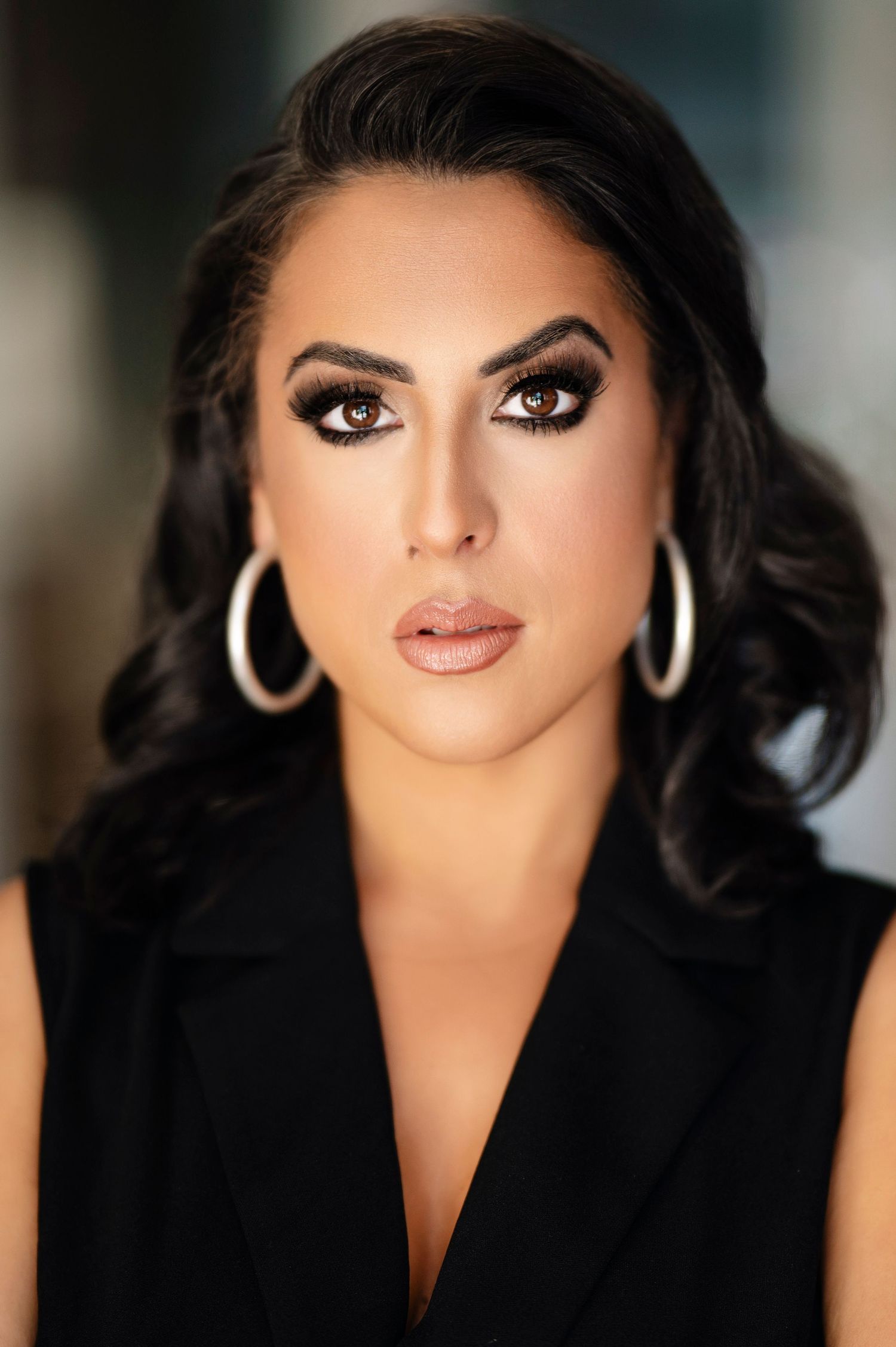 A glamorous portrait featuring dark wavy hair, dramatic eye makeup, hoop earrings, and a black sleeveless top against a soft background.