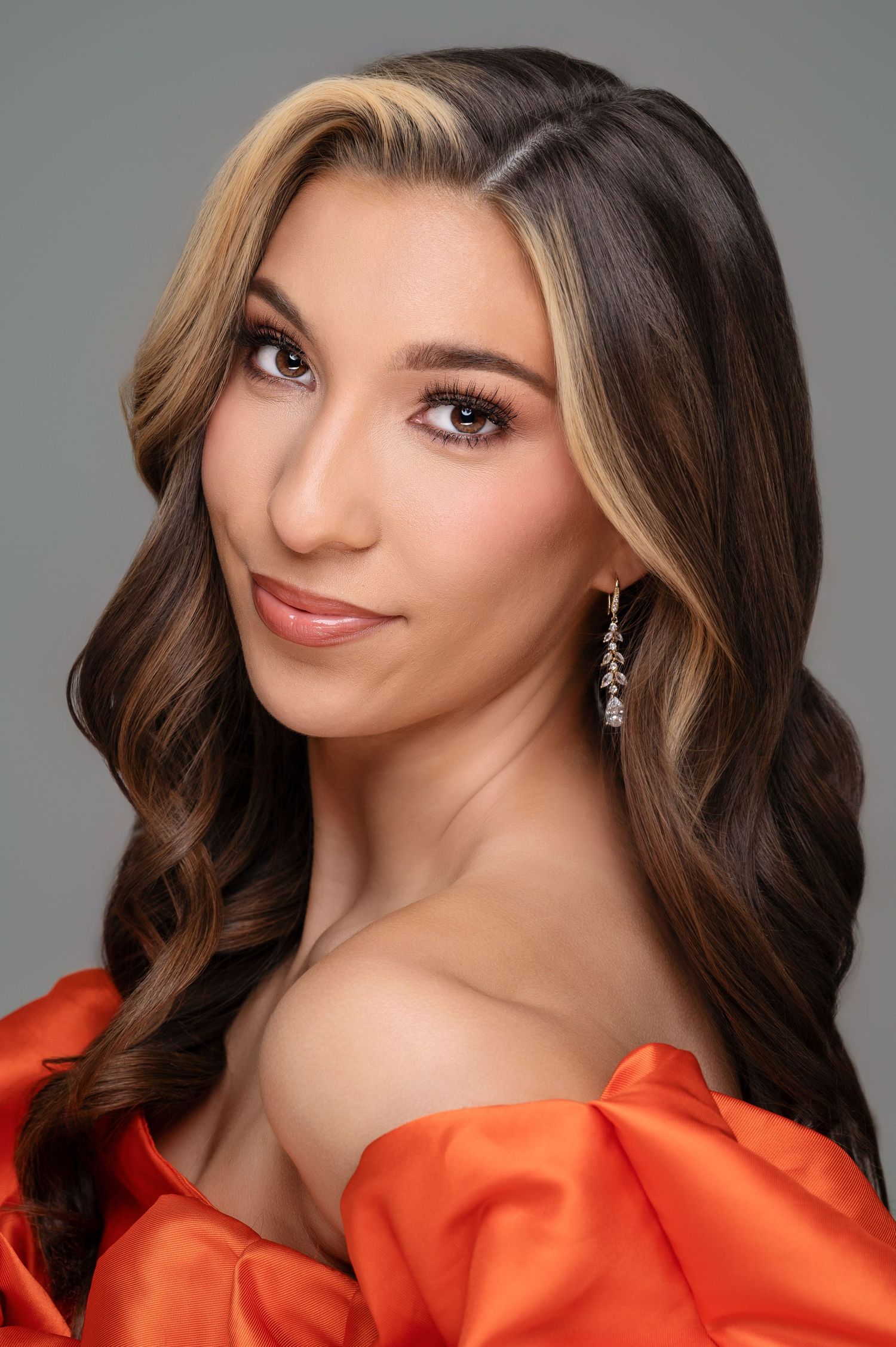 A woman with flowing brown hair and dangling earrings poses in a bright orange off-shoulder dress against a grey backdrop.