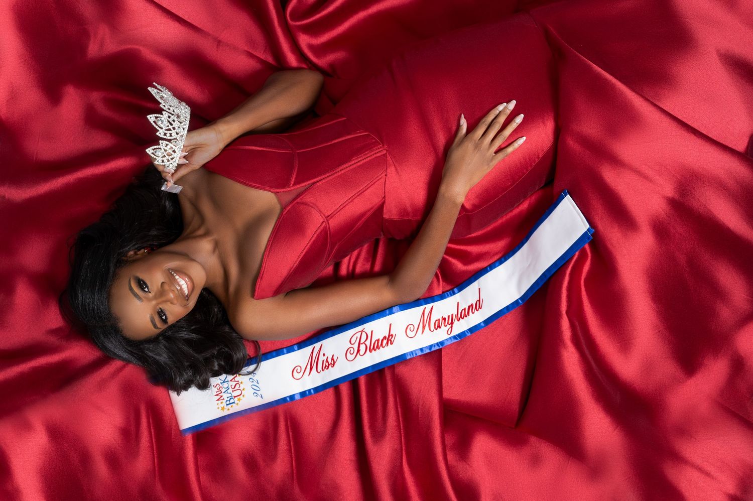 A person in a red satin dress lying on red satin fabric with a white and blue sash across their body.