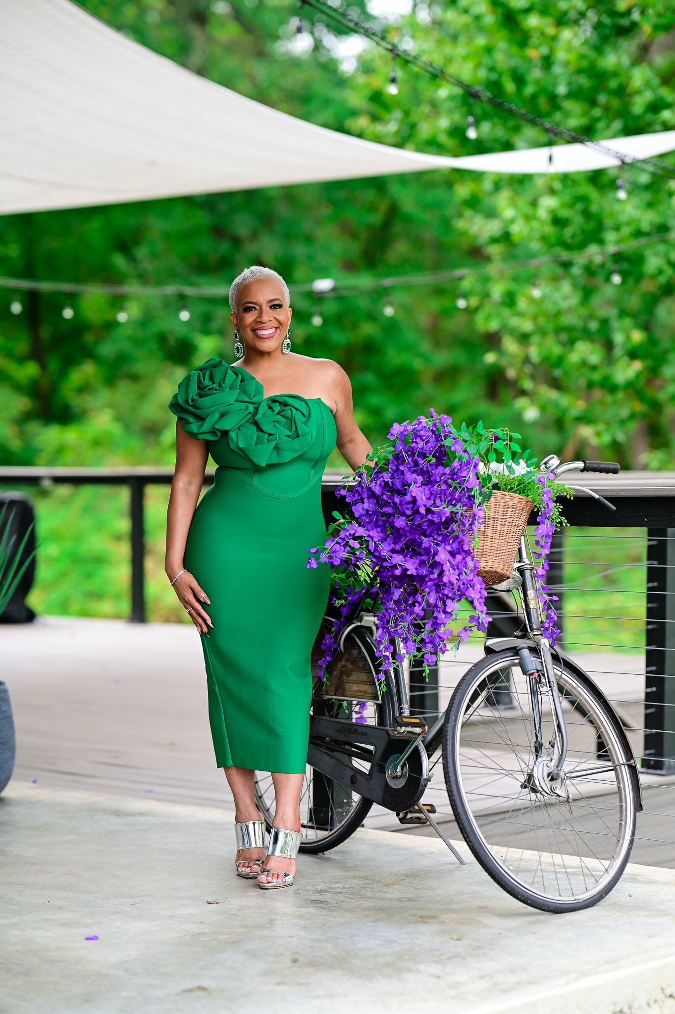 Woman in green one-shoulder ruffle dress posing beside a bicycle adorned with purple flowers on an outdoor patio.