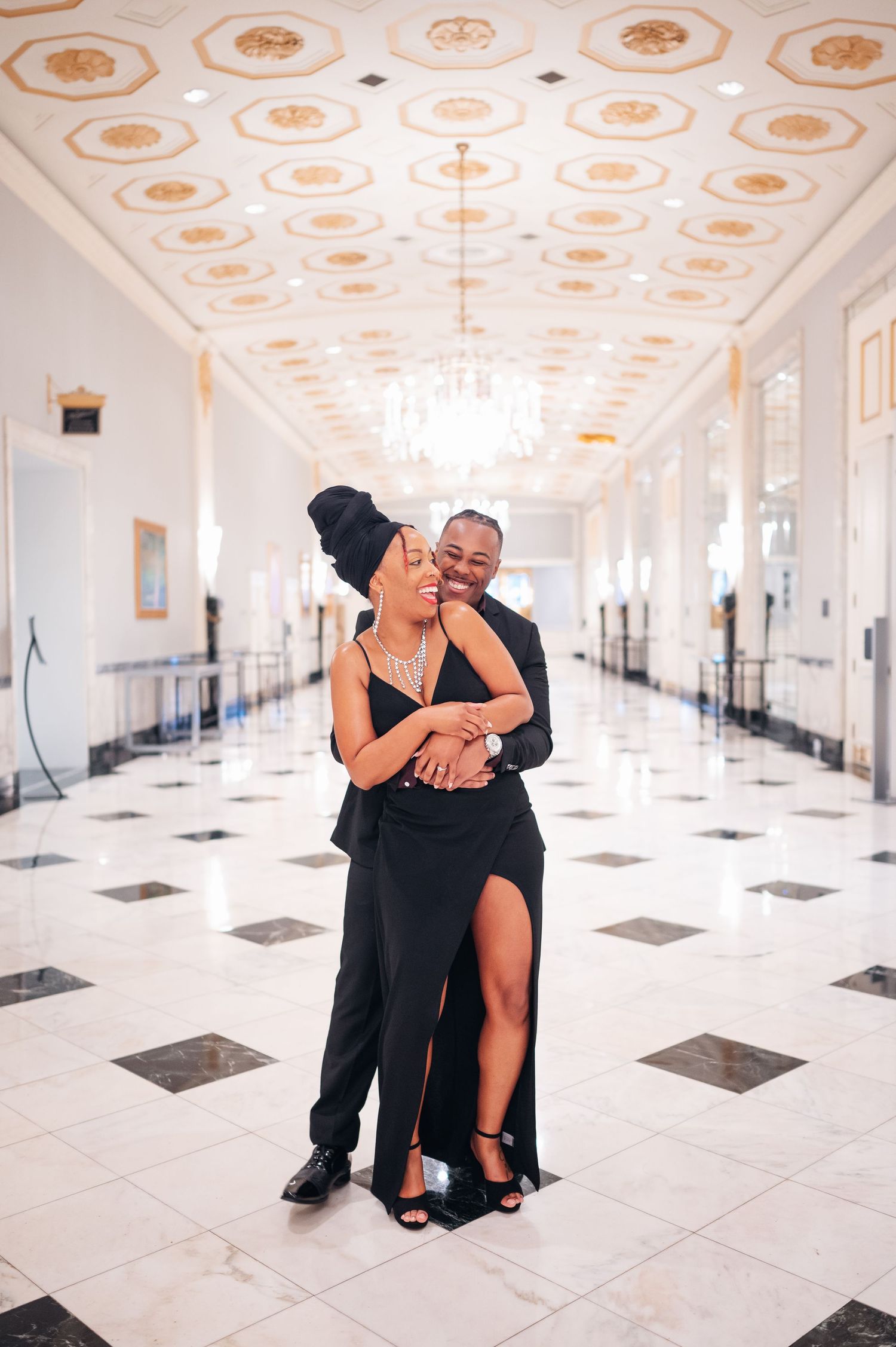 A couple embraces in a grand hallway with ornate ceiling details, black and white checkered floor, and a crystal chandelier.