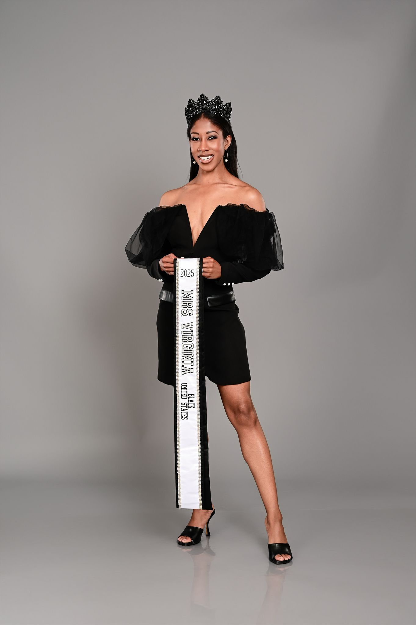 A woman in a black mini dress and long cardigan poses confidently against a gray studio backdrop.