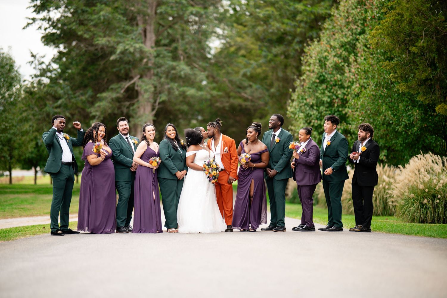 Wedding party members stand in a line formation on a green lawn surrounded by trees during an outdoor ceremony.