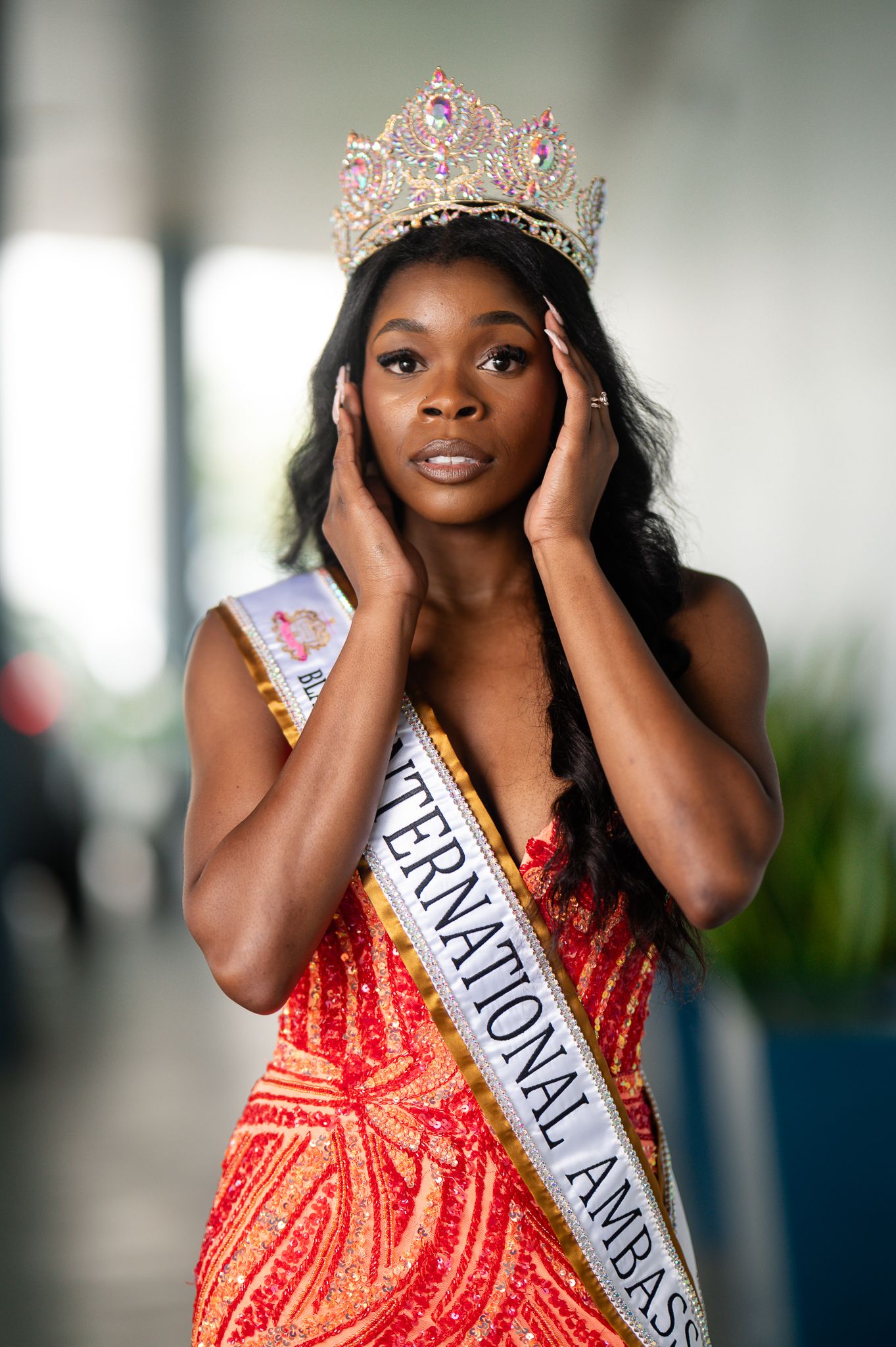 A pageant contestant wearing a sparkling crown and red sequined gown poses with hands framing their face.