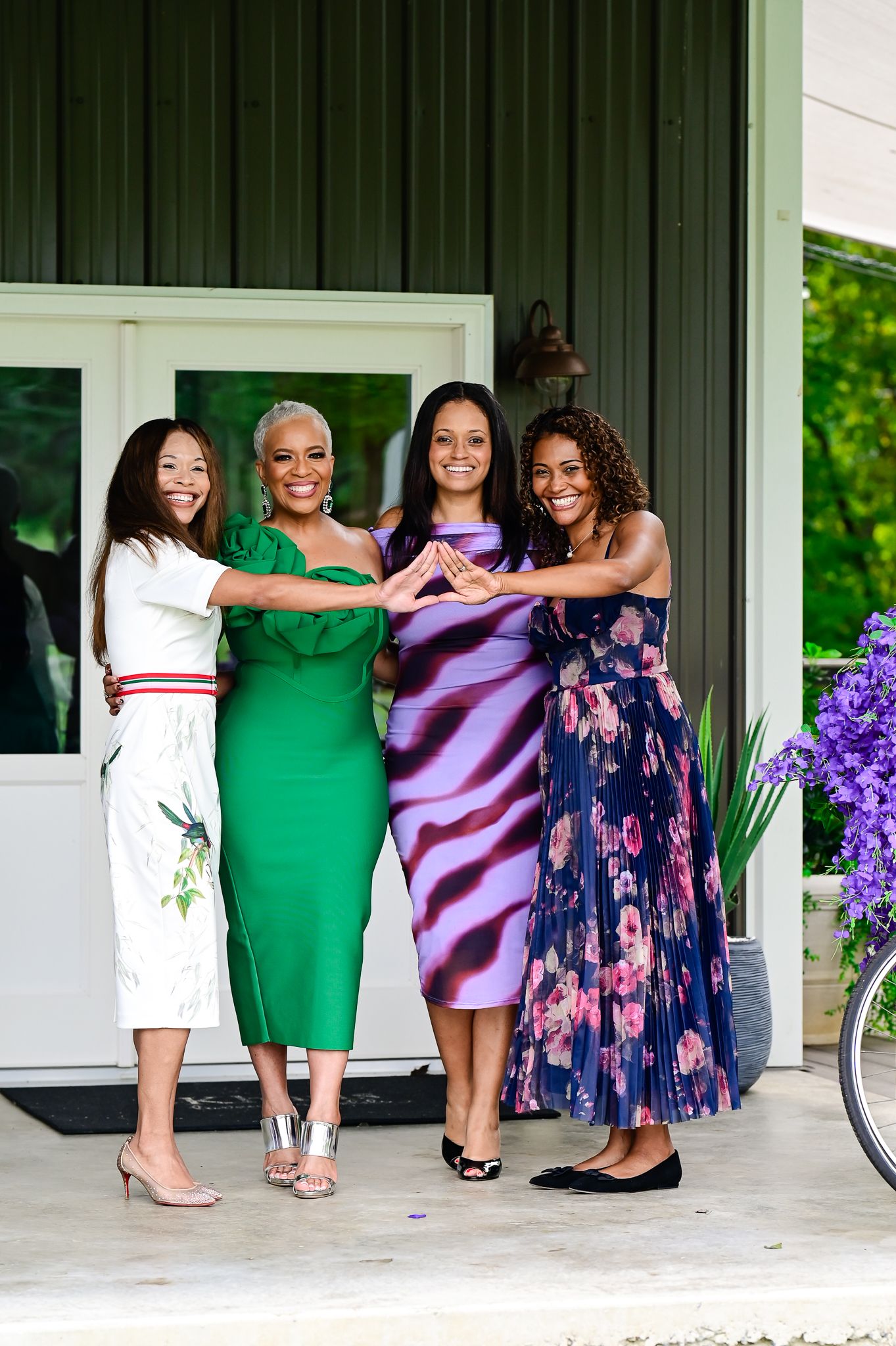 Four women in colorful dresses smiling and posing together on a porch, forming a heart shape with their hands.