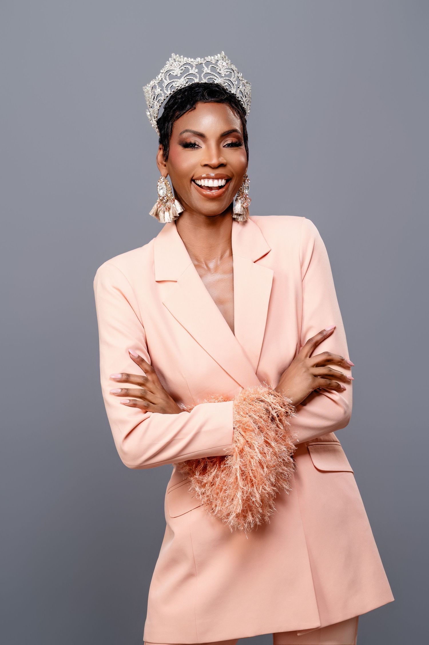 A person wearing a sparkling crown and peach blazer with feathered cuffs poses against a gray background.