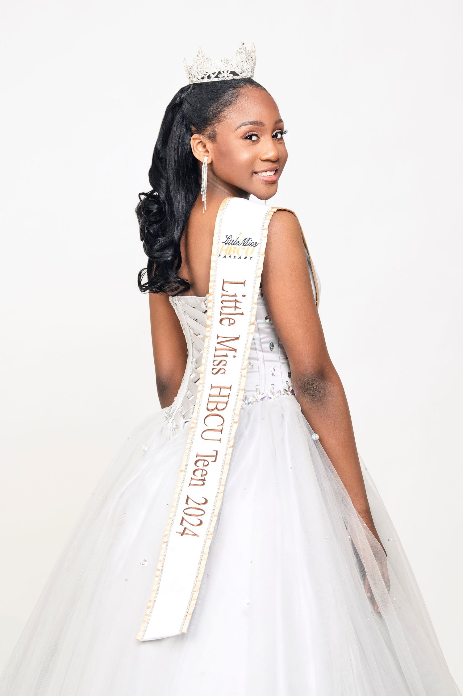 A pageant contestant wearing a white ball gown and crown poses elegantly with a sash against a light background.