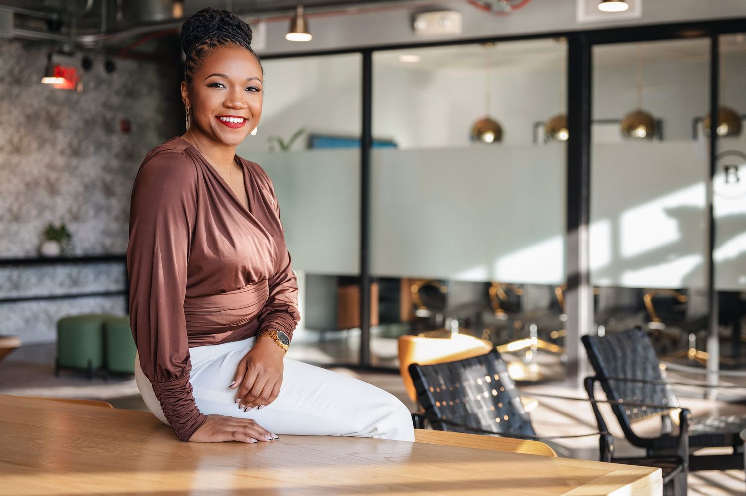 Professional portrait of someone in brown blouse and white pants sitting casually on office desk in modern workspace.