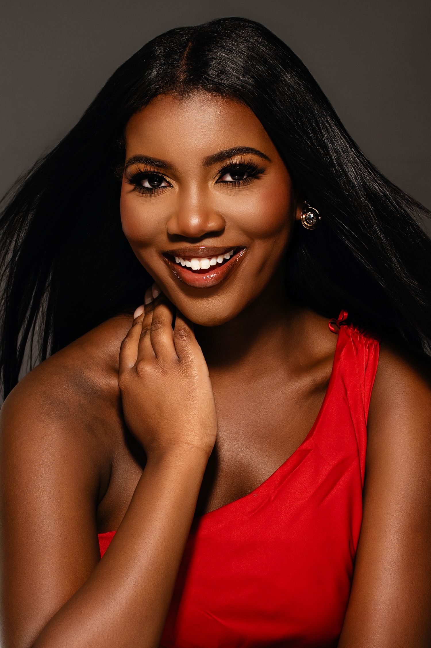 Smiling young Black woman with long straight hair wearing a red one-shoulder top, posing confidently against a gray background.