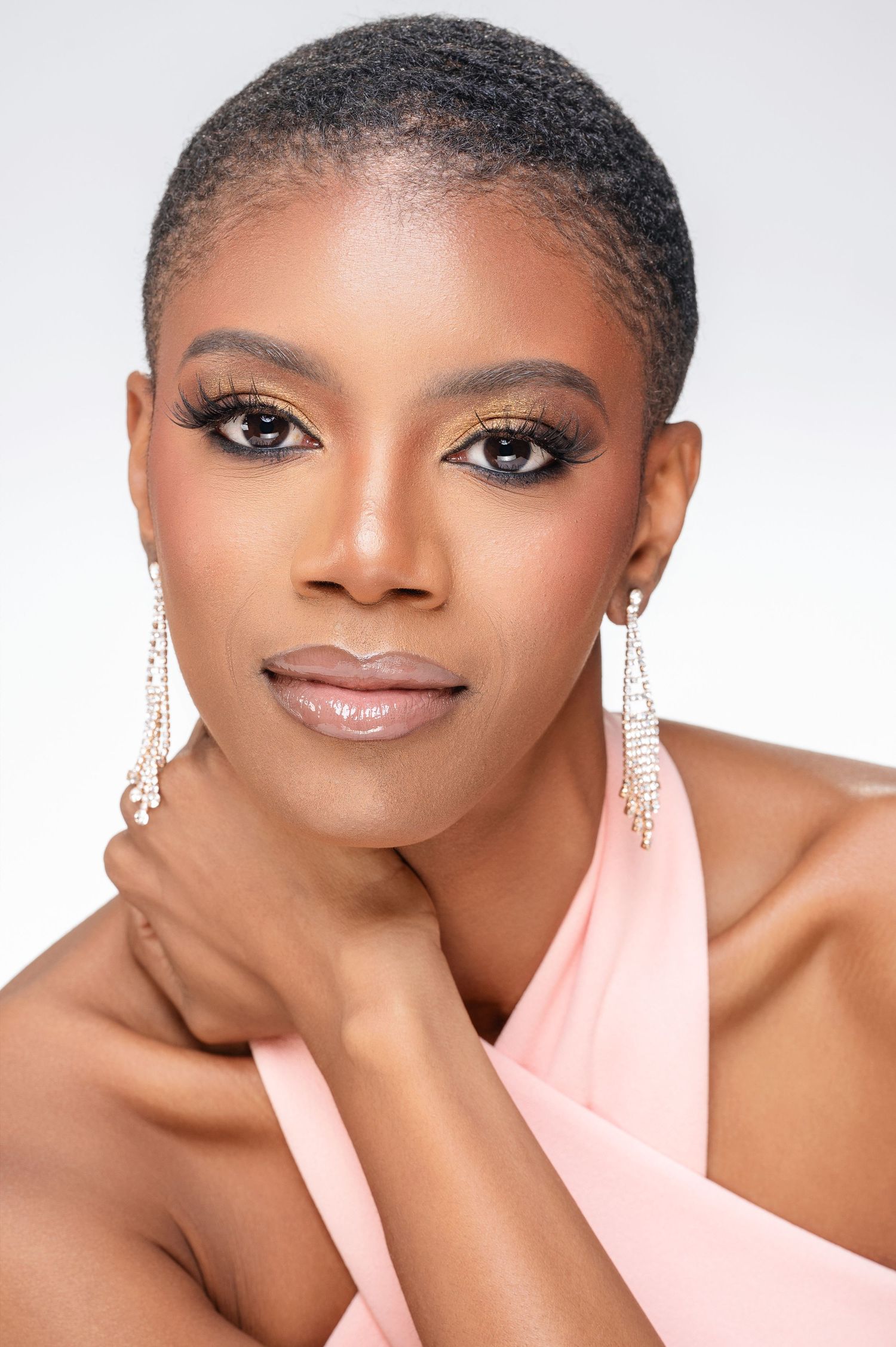 A glamour portrait of someone in soft pink fabric wearing dazzling crystal earrings with dramatic eye makeup and a warm smile.
