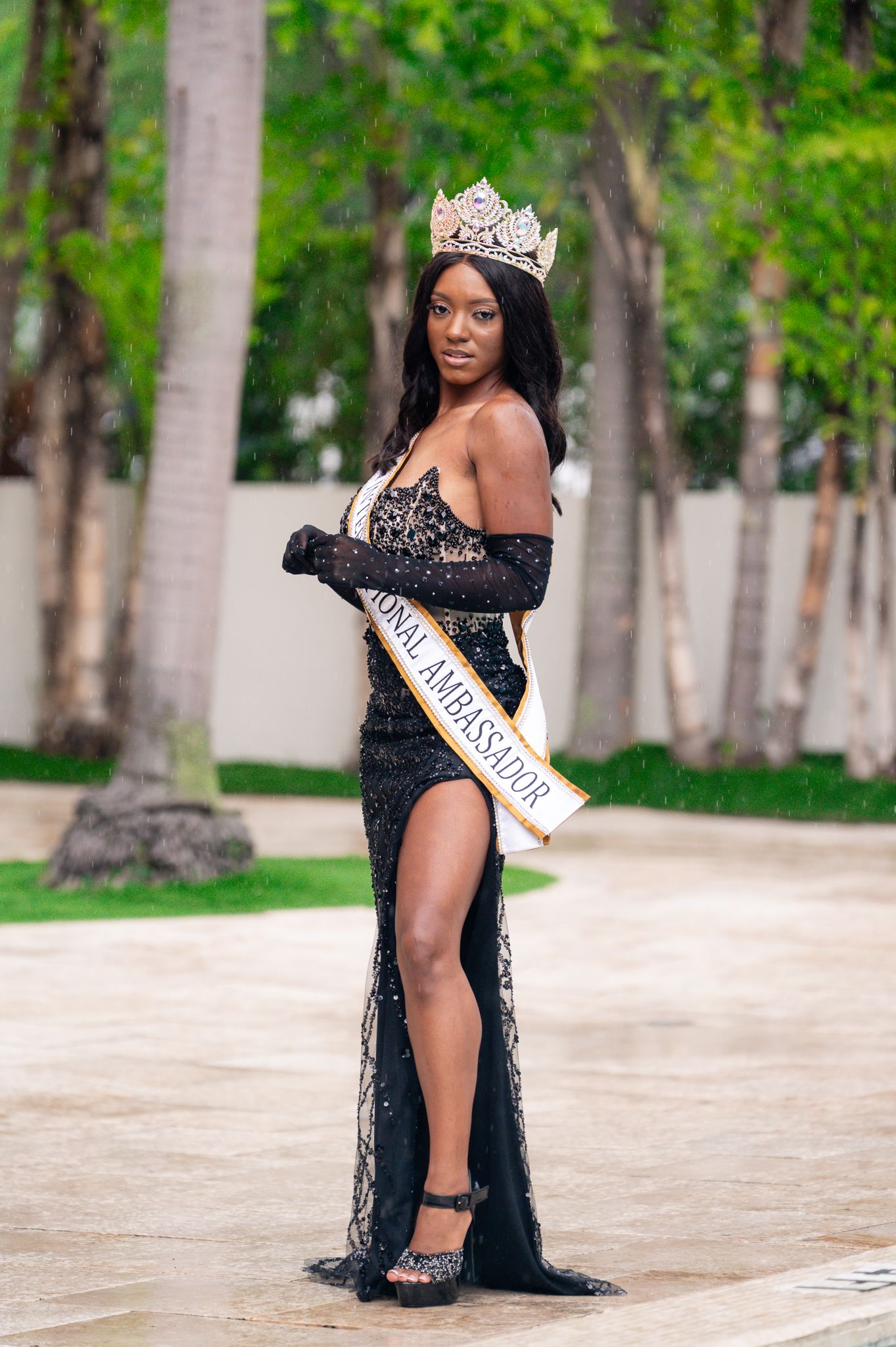 A pageant winner poses in an elegant black lace gown with a thigh-high slit and crown in an outdoor setting.