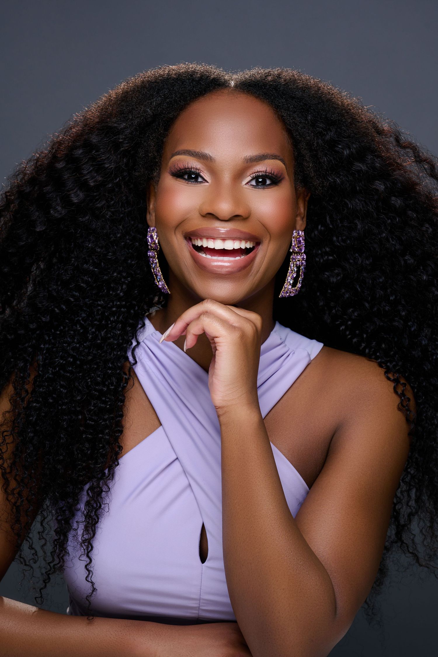 Portrait of a joyful woman with natural curly hair wearing a lavender top and sparkly earrings against dark background.