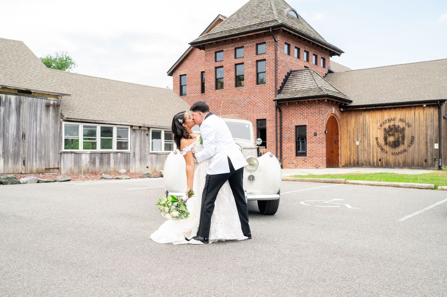 A couple in wedding attire share a romantic moment in a parking lot beside a vintage car near a brick building.