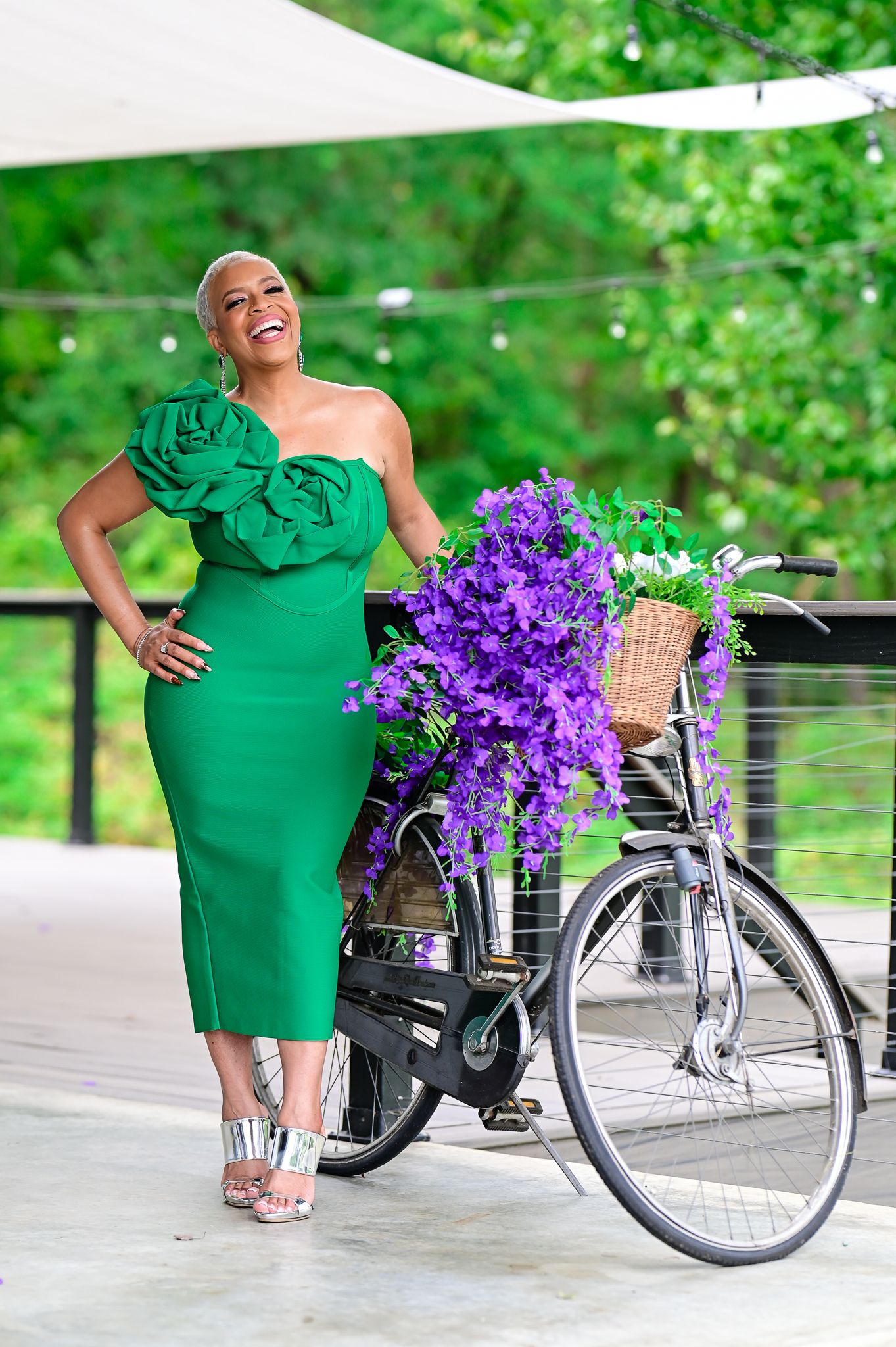 Woman in green strapless dress poses beside a bicycle adorned with purple flowers outdoors.