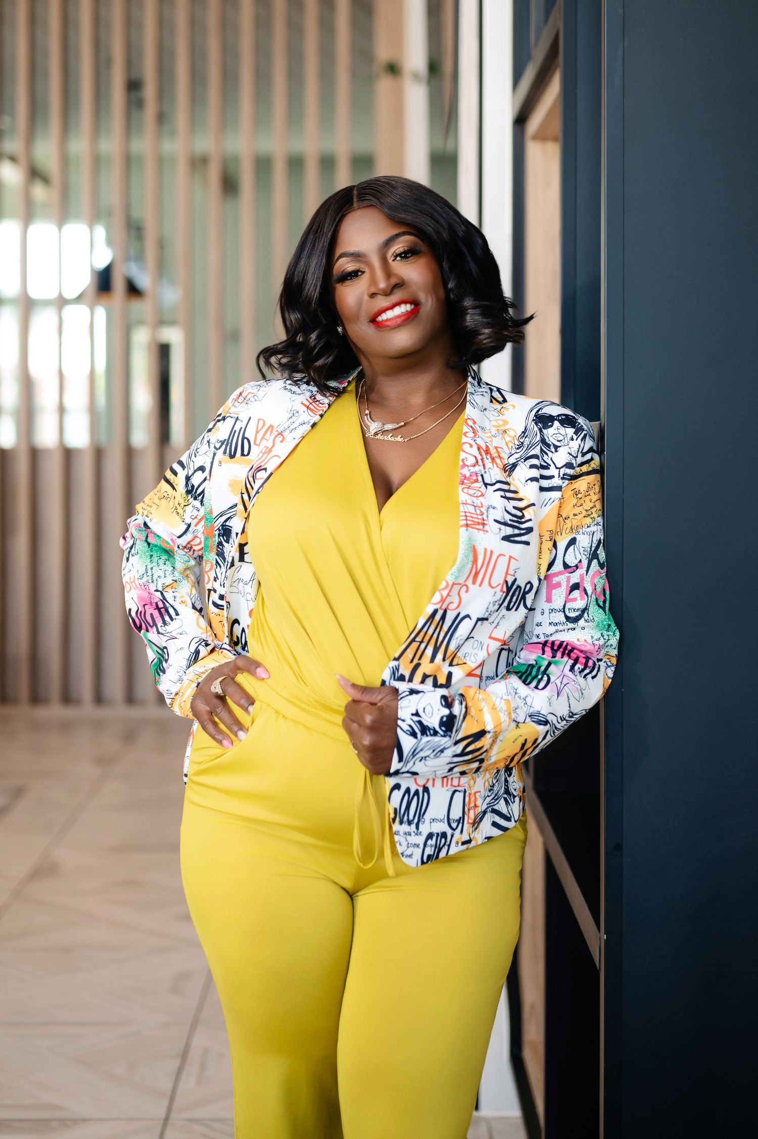 Woman in yellow jumpsuit and colorful graphic blazer posing confidently against a modern building exterior.