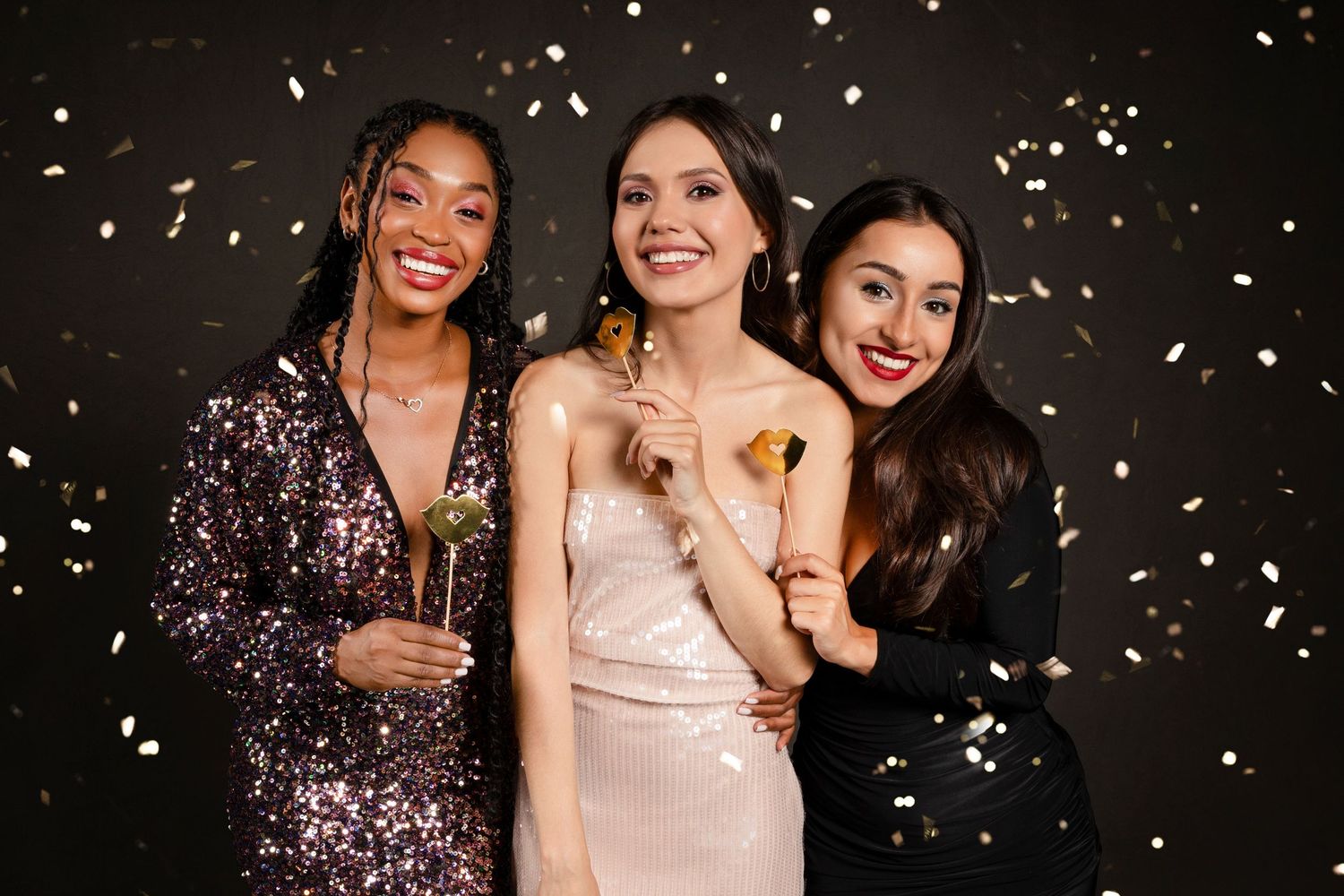 Three elegant women in party dresses celebrate with champagne glasses and falling gold confetti on black background.